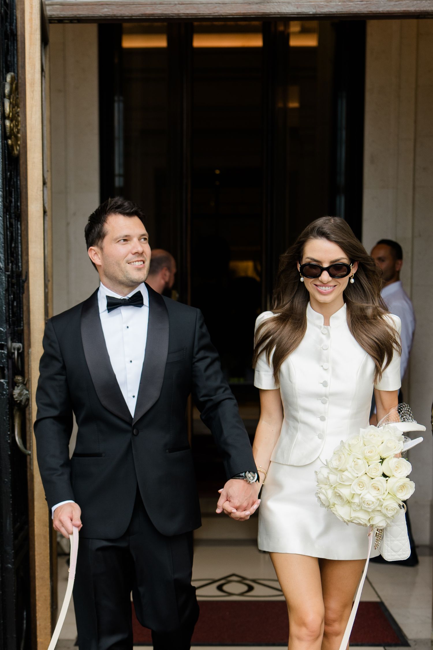 Couple exits in wedding attire, groom in tuxedo and bride in white mini dress with bouquet, holding hands.
