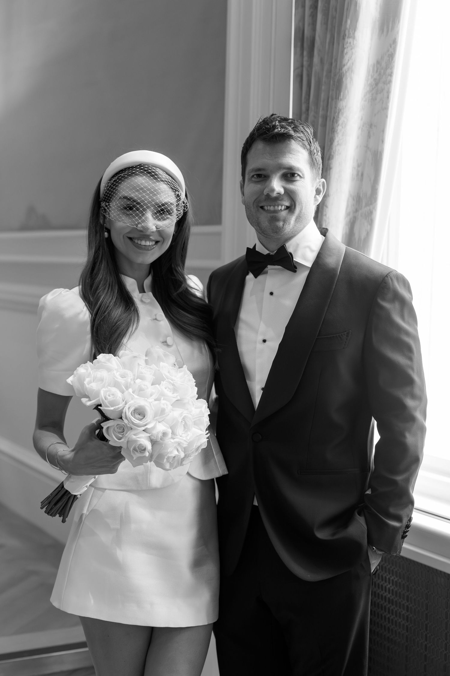 Black and white photo of a smiling bride in mini dress with veil bouquet and groom in tuxedo posing together.