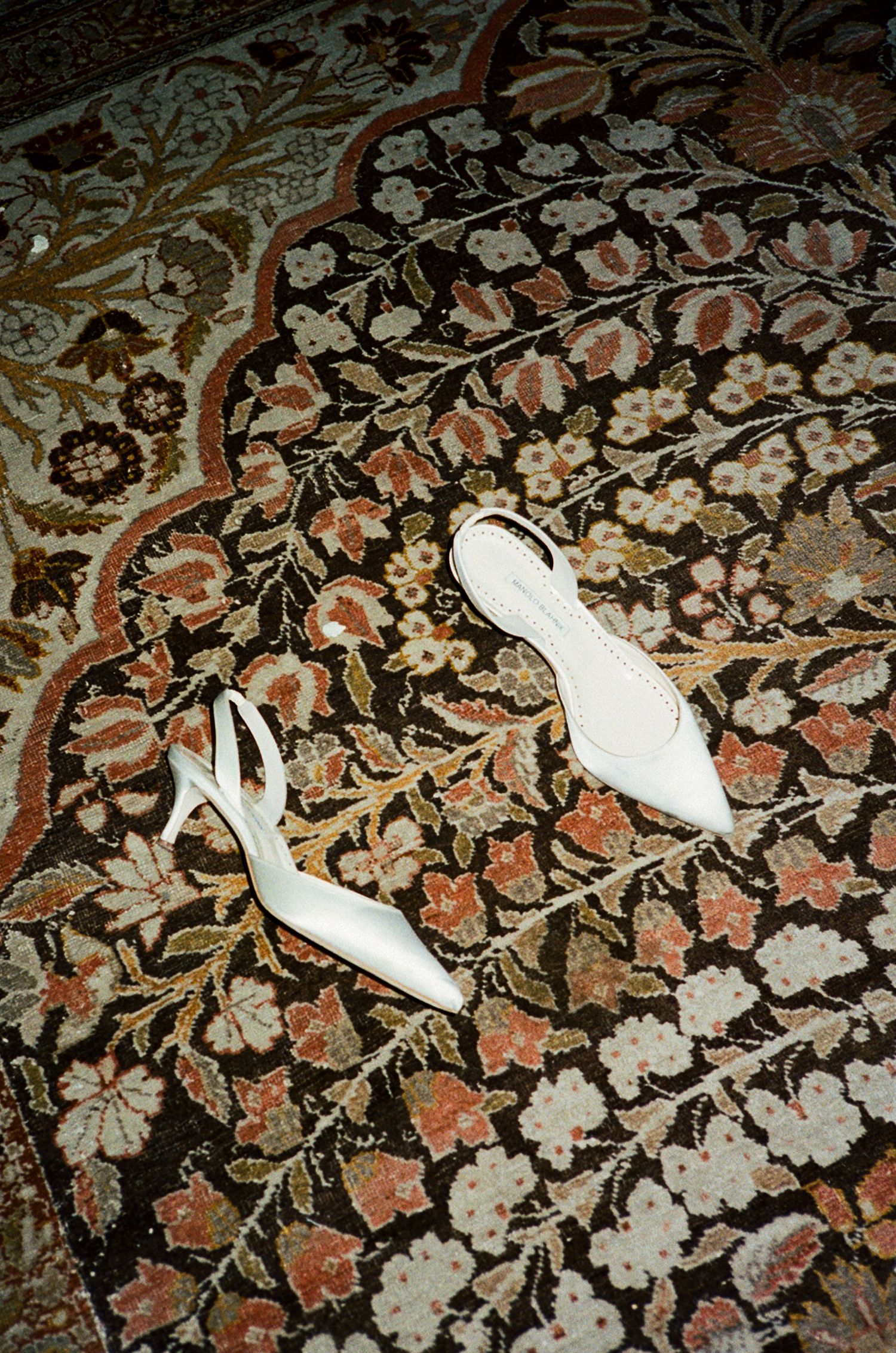 White slingback heels displayed on an ornate floral patterned rug with rich red and gold tones.