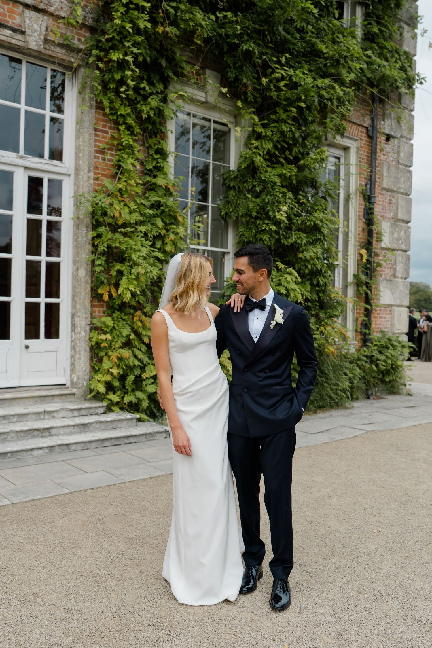 Bride and groom posing before ivy-covered stone manor house in elegant wedding attire.