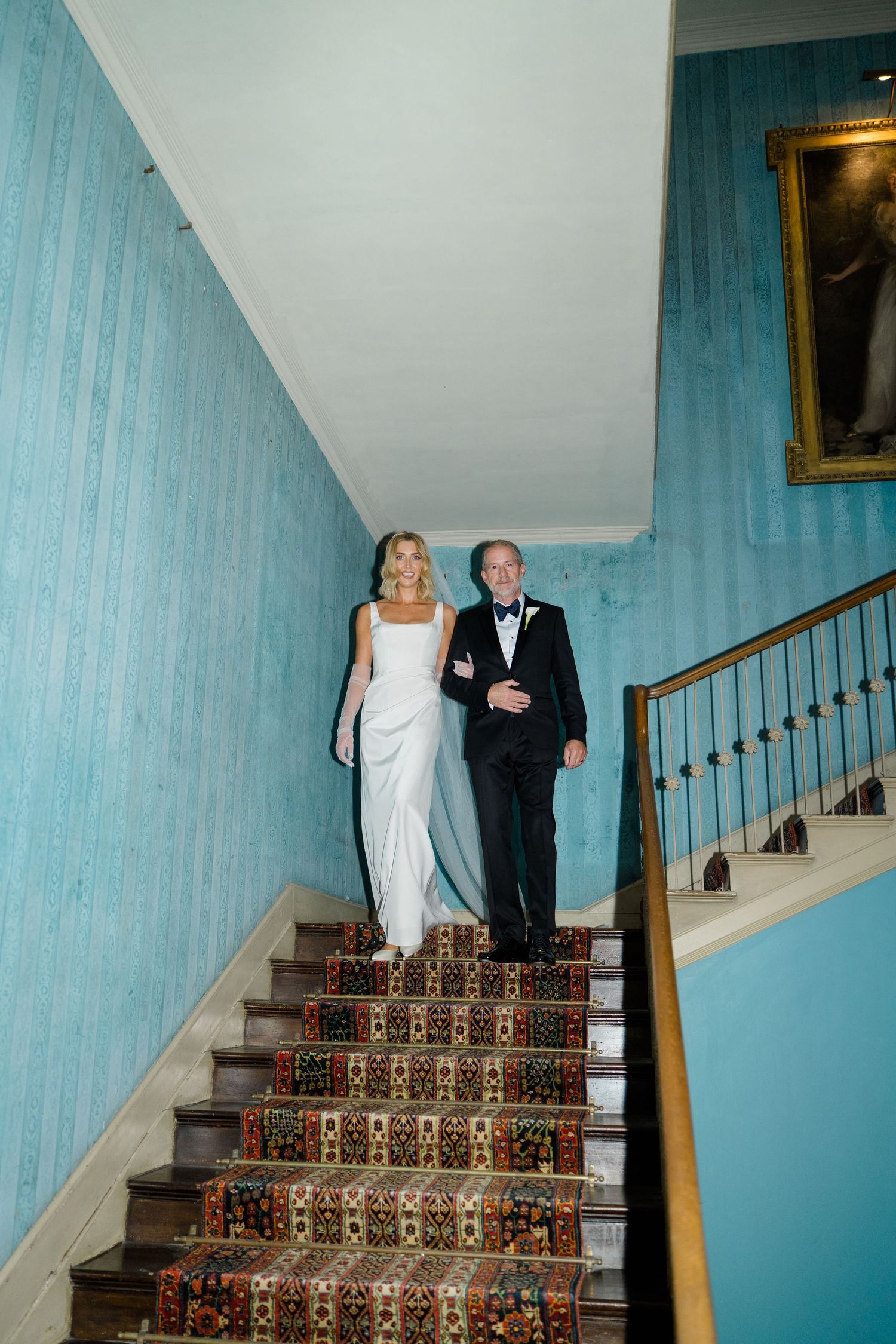 Bride in white gown descending ornate staircase with older gentleman in tuxedo at wedding venue.