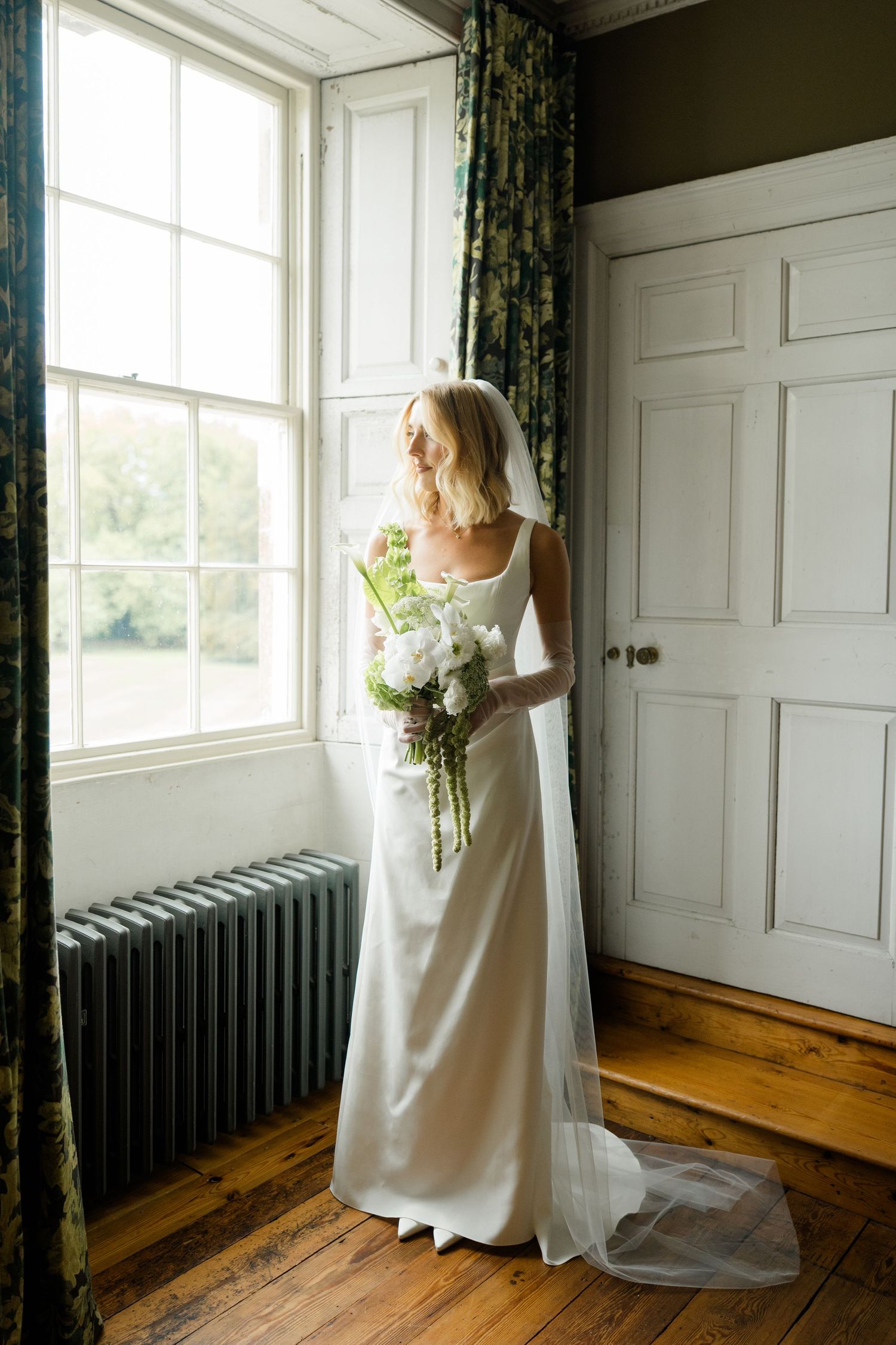 Bride in white satin gown with veil holding bouquet while gazing out tall window.