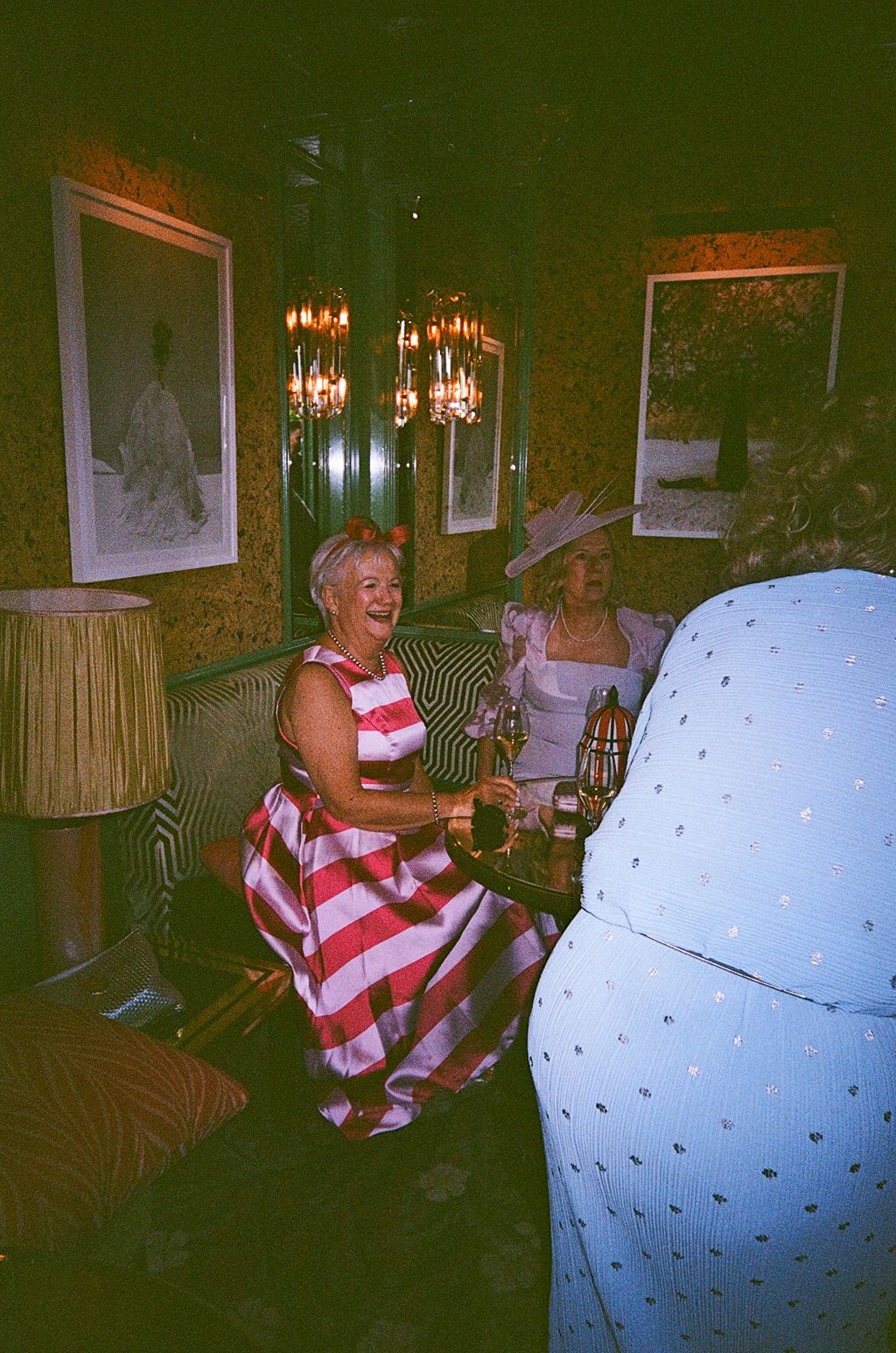Elegantly dressed women laugh together in a richly decorated room with chandelier lighting.