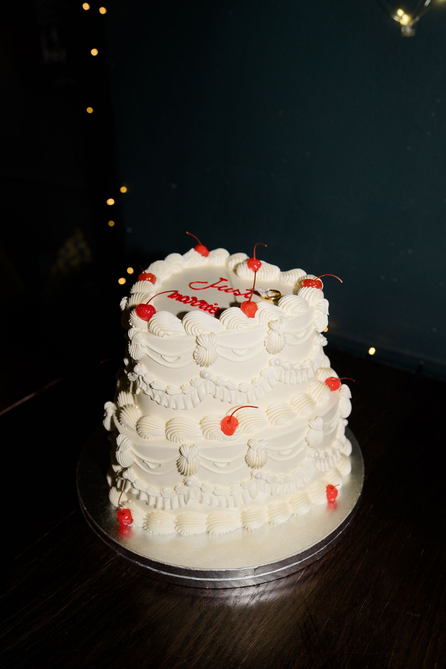 Two-tier white wedding cake decorated with red cherries and piped cream frosting with bokeh lights.