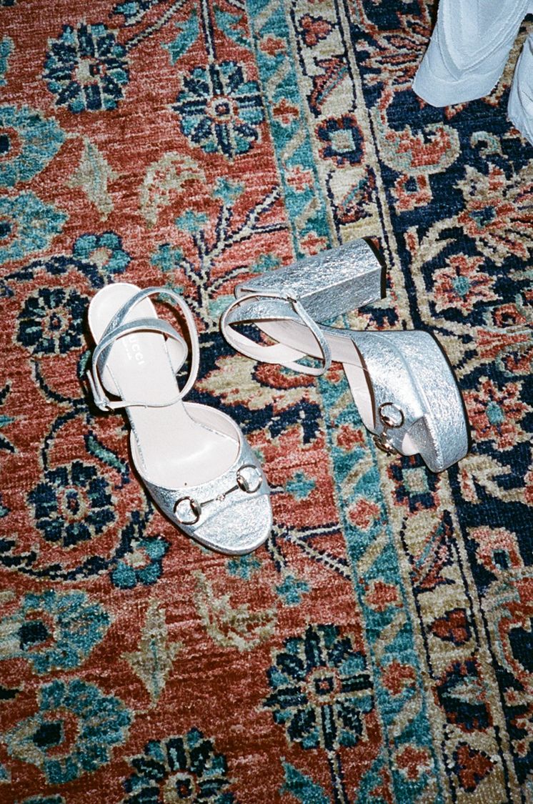 Silver metallic block heel sandals with ankle strap and gold hardware, displayed on a vintage red floral patterned rug.