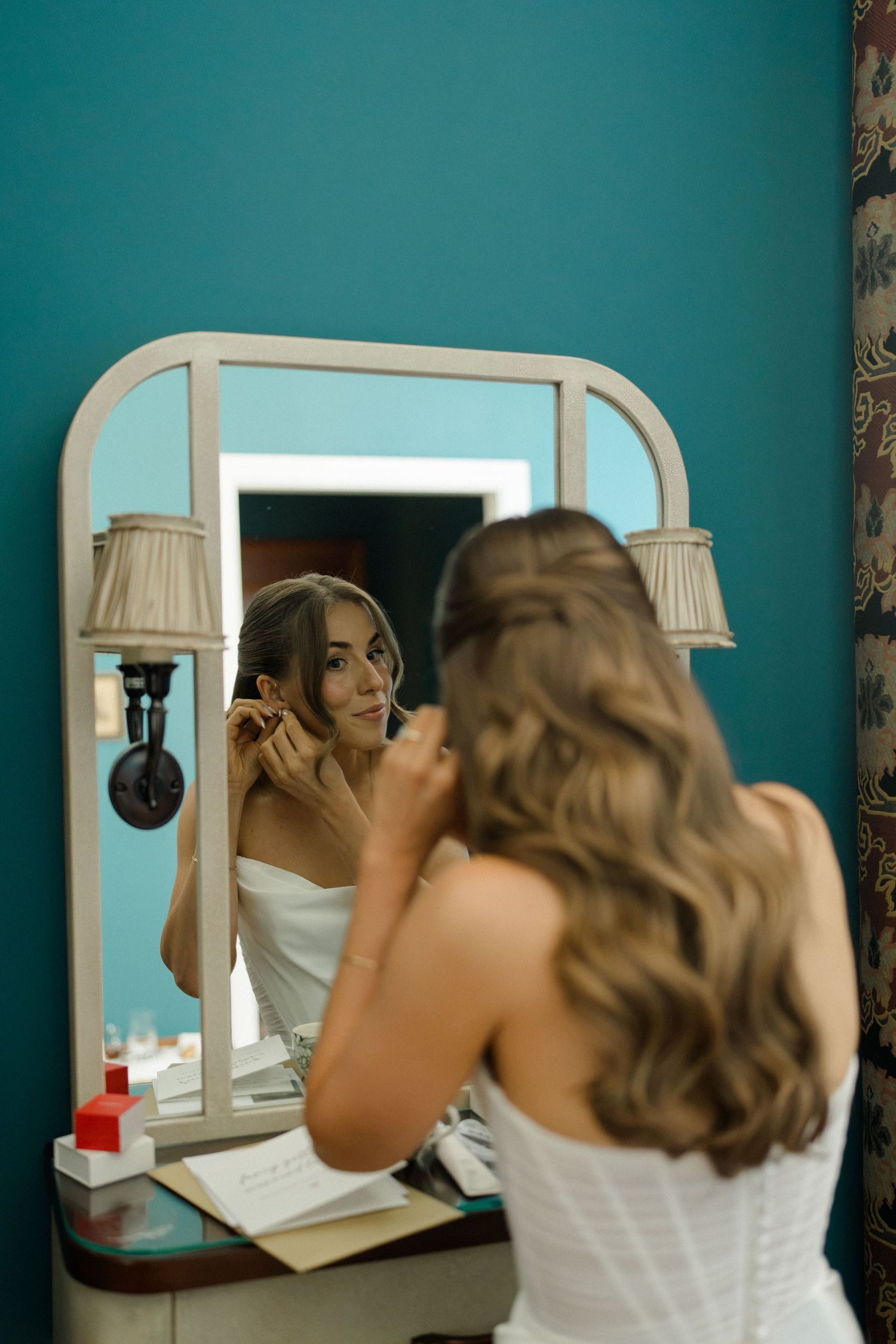 Bride applying makeup at a vintage vanity mirror with lights in a teal-walled dressing room.