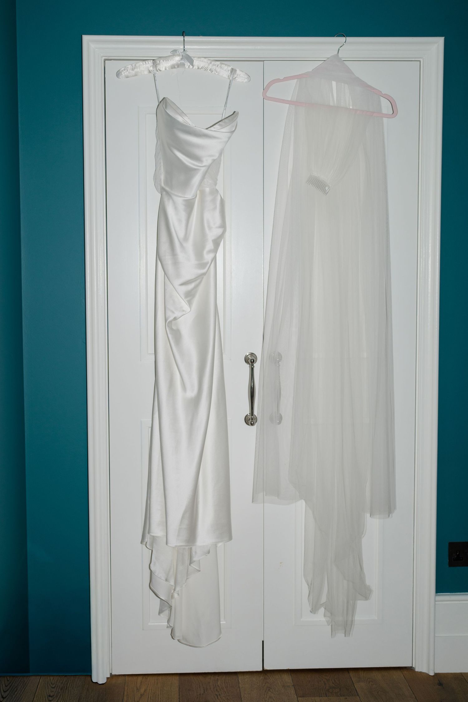 White satin wedding dress and sheer veil hanging on a white door against a teal wall.
