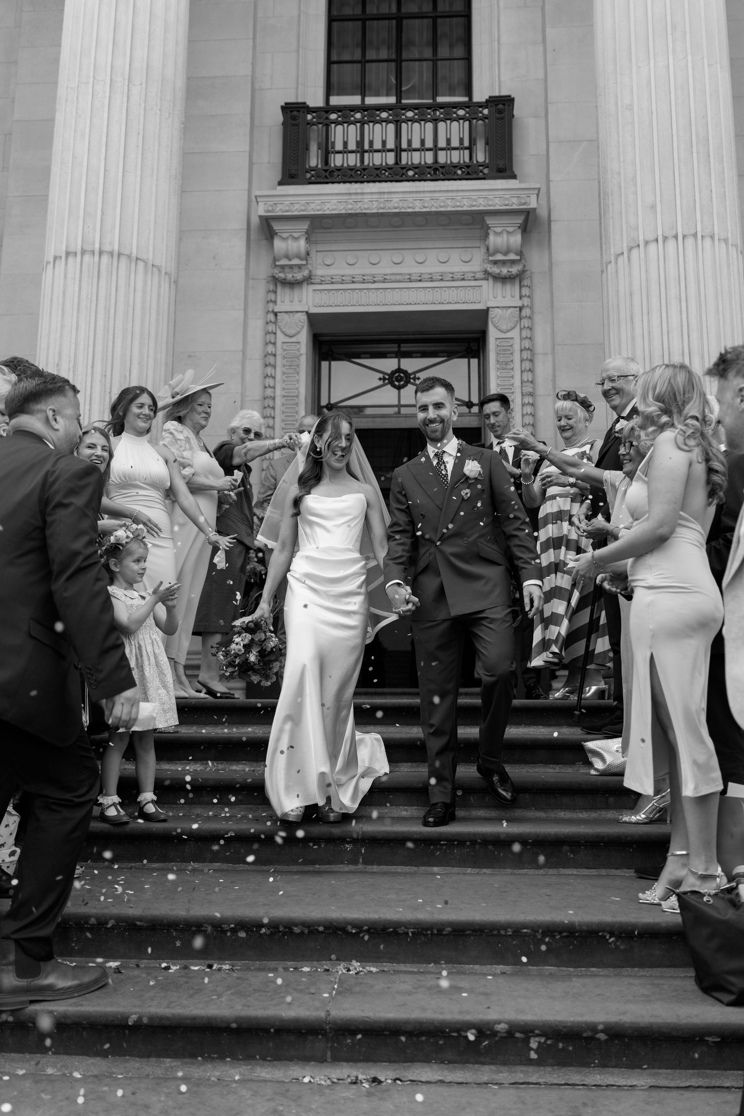 Black and white photo of newlyweds descending steps through confetti shower with cheering wedding guests.