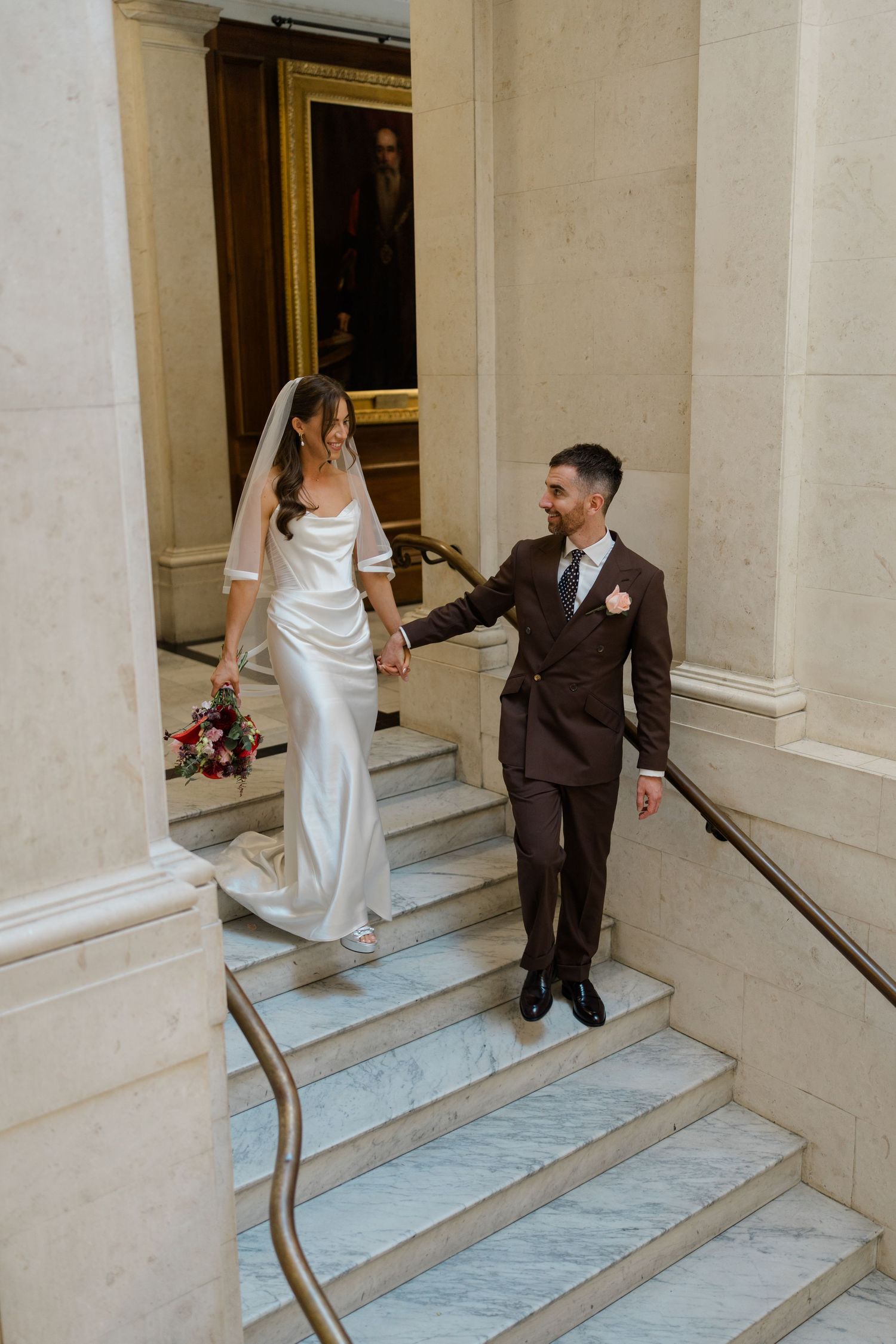 Bride and groom hold hands while walking up marble stairs at their elegant wedding venue.