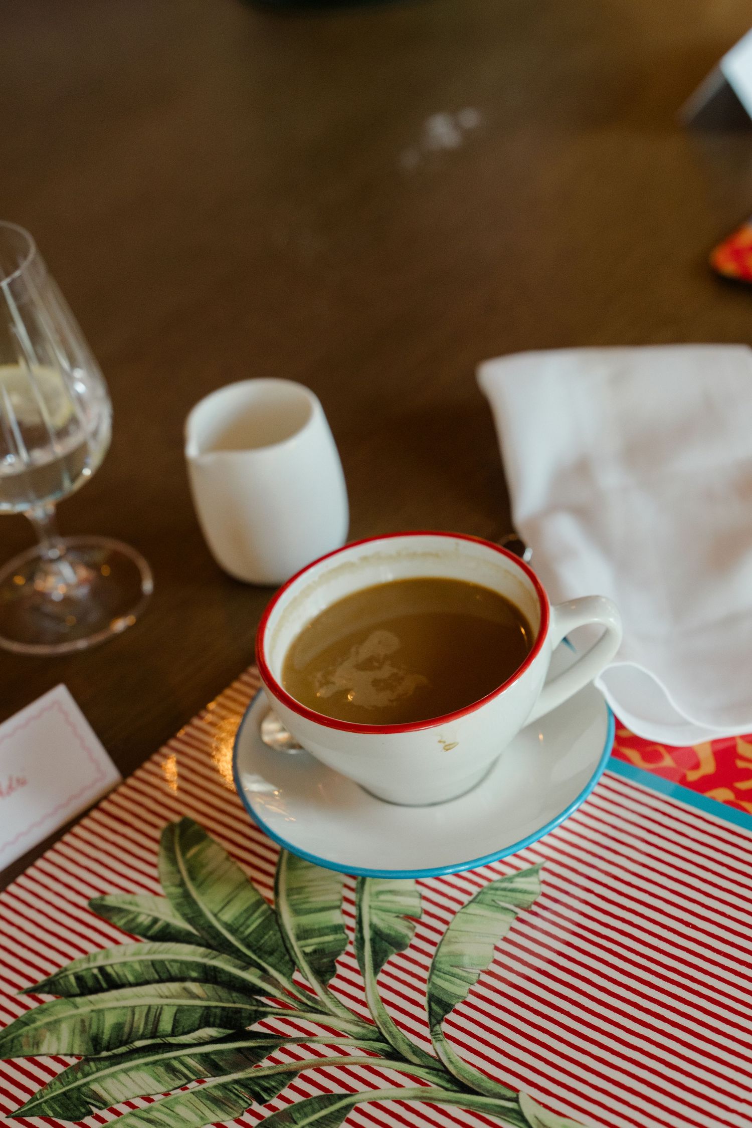 White cup of black coffee with red rim on tropical leaf placemat at a wedding reception table.
