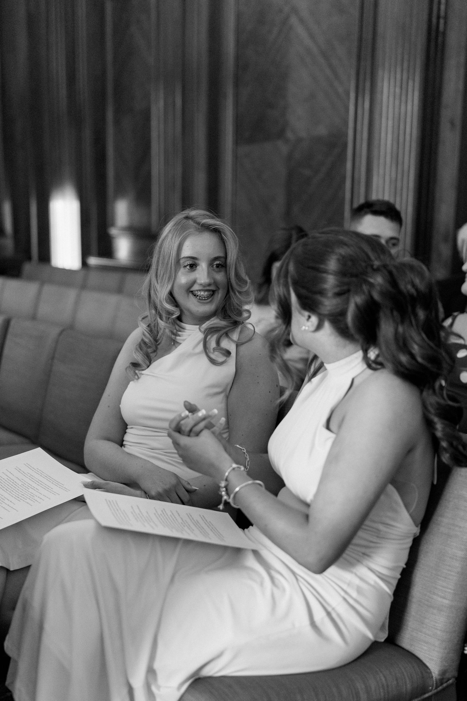 Black and white photo of two bridesmaids in white dresses chatting and smiling during wedding ceremony.