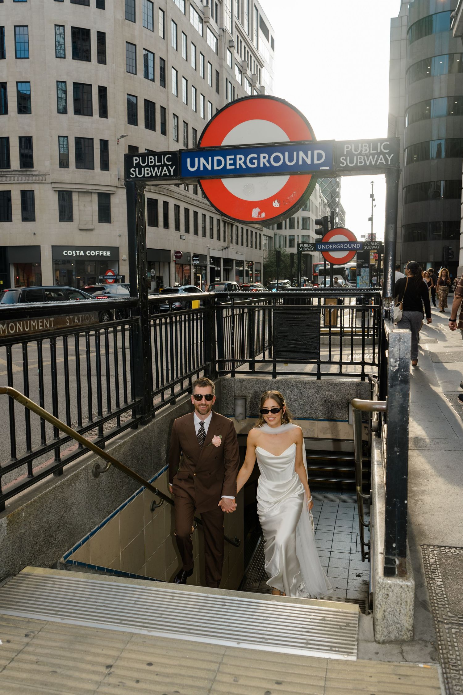 Newlyweds in wedding attire emerging from London Underground Monument station entrance.