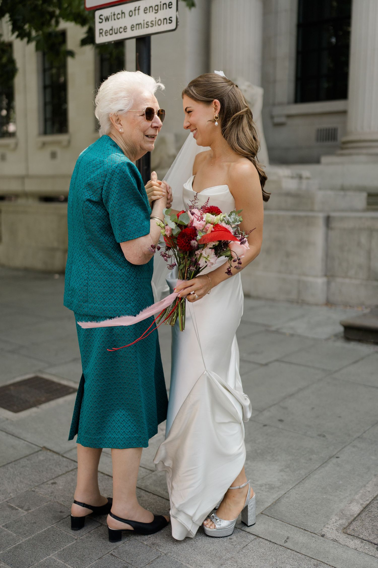 Elderly woman in teal dress smiles with bride holding red bouquet outside London registry office.