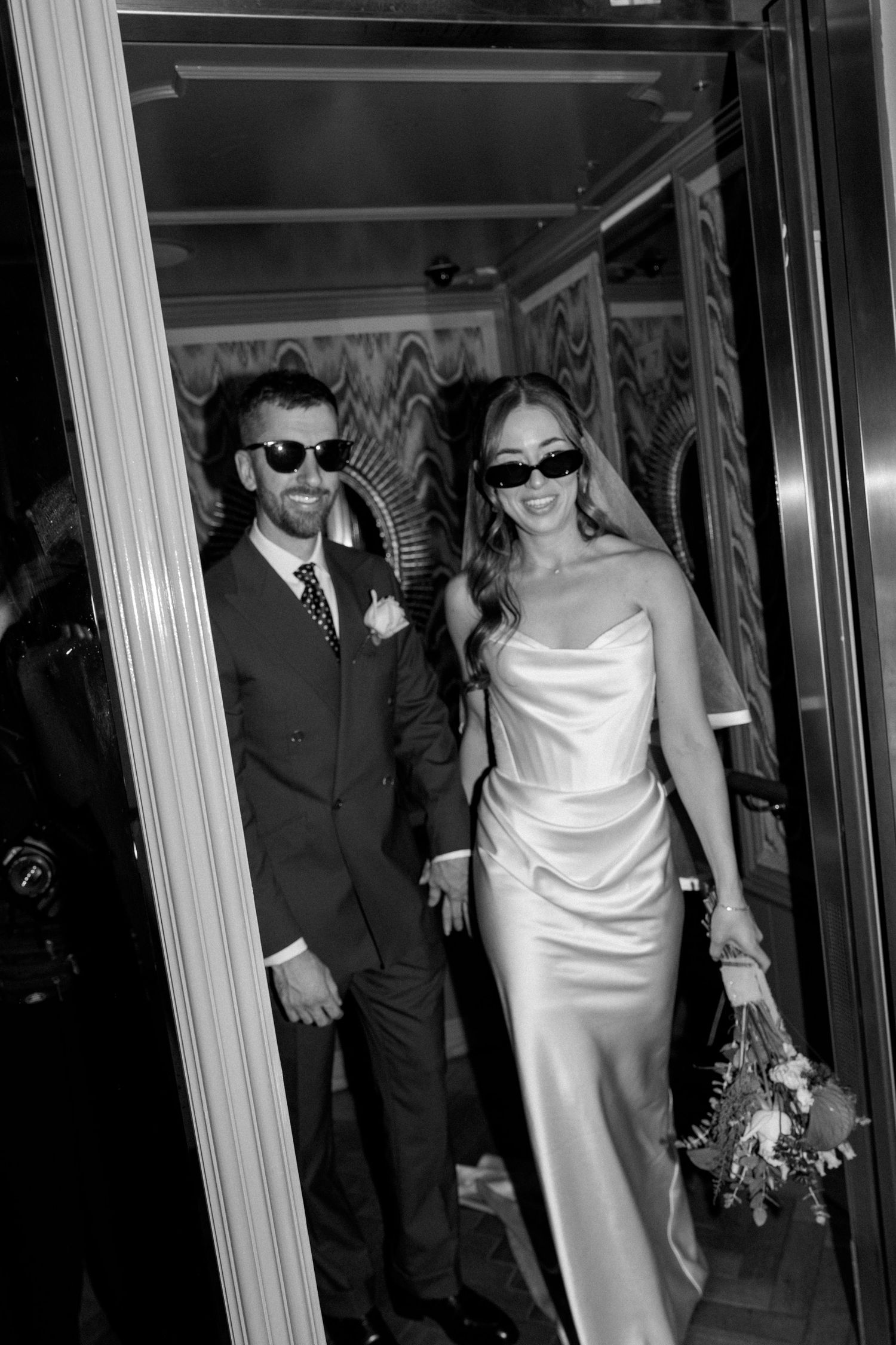 Stylish bride and groom wearing sunglasses in black and white wedding portrait indoors.