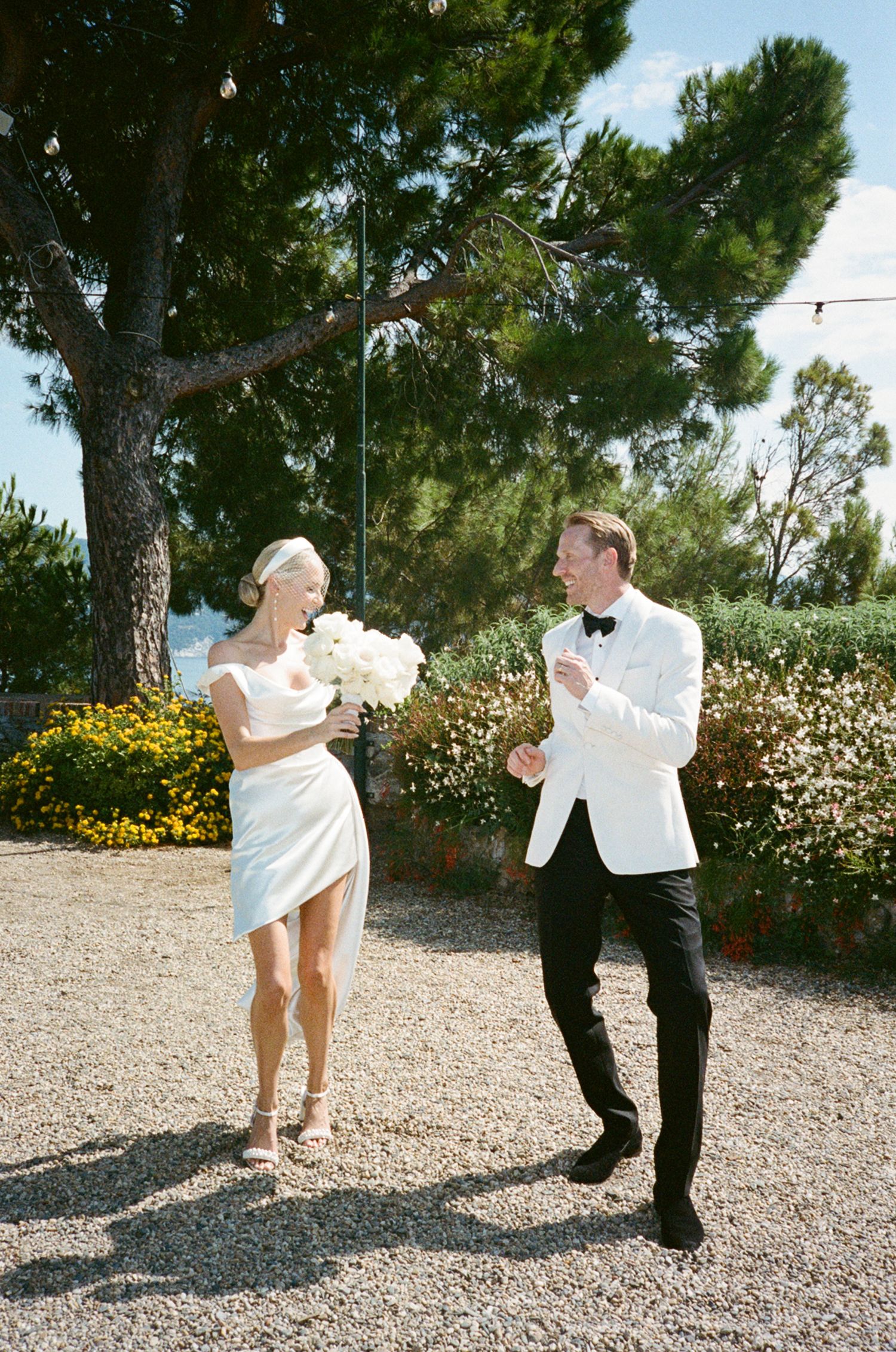 Laughing bride with white bouquet and groom in white tuxedo celebrate on a sunlit gravel path in Italy.