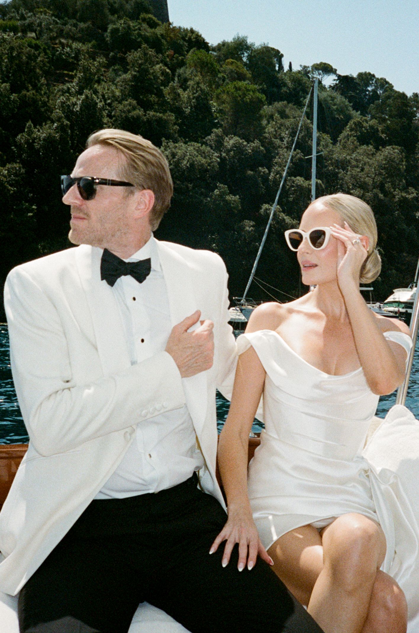 Elegant couple in white wedding attire wearing sunglasses seated on a luxury boat in a Mediterranean harbor.