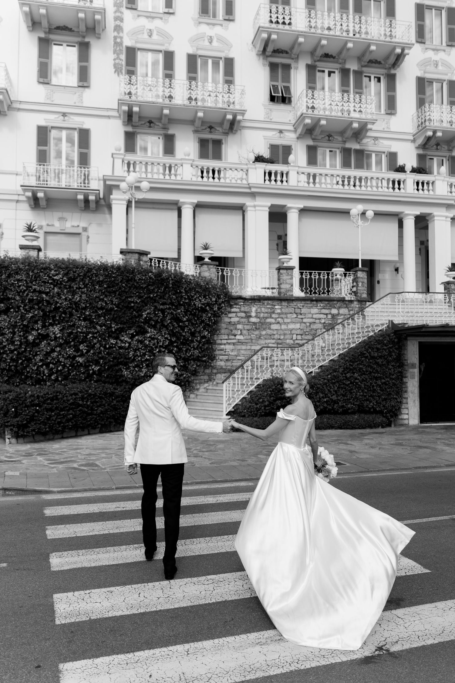 Bride and groom crossing zebra crossing in black and white photo outside grand European hotel.