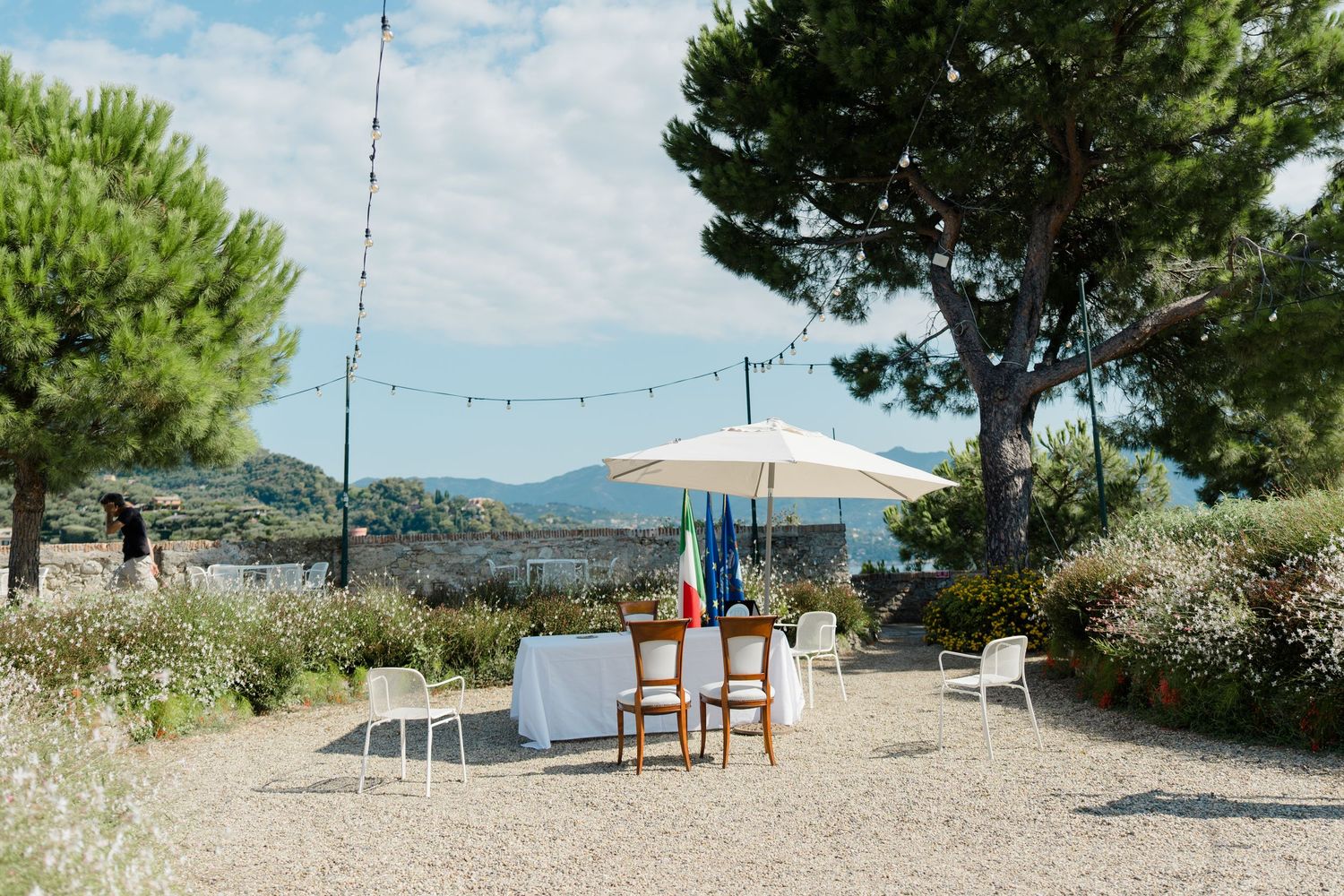 Outdoor wedding ceremony setup with white umbrella, table, and chairs on gravel surrounded by lush greenery and string lights.
