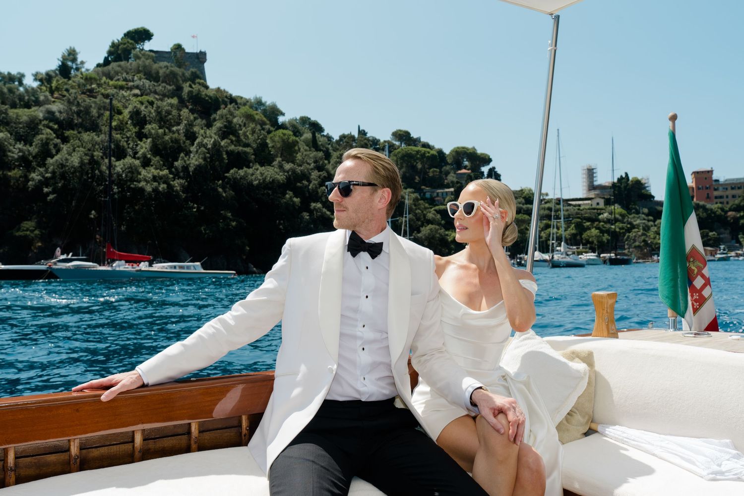 Elegant couple in wedding attire relaxing on classic wooden boat on sparkling blue Italian waters.