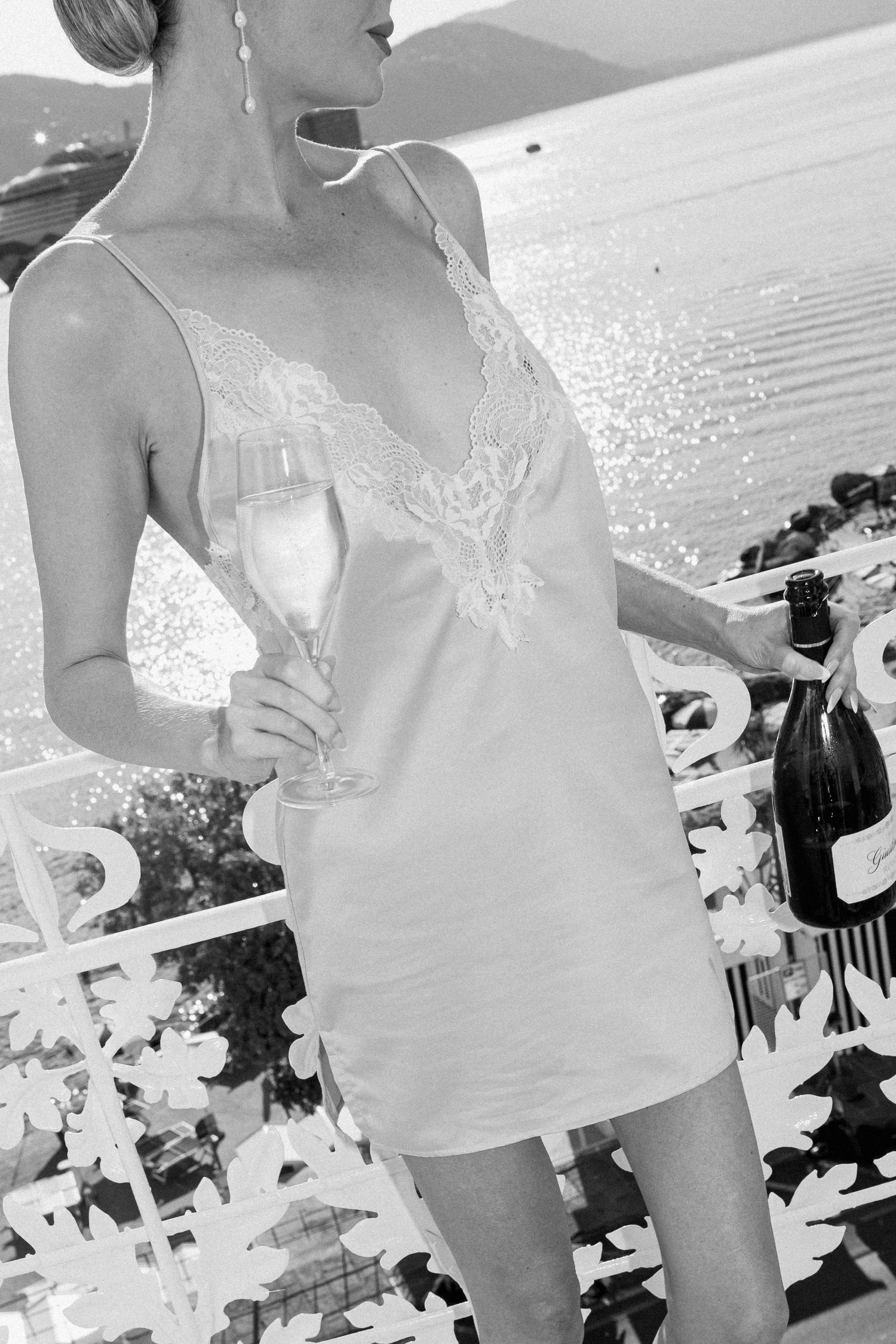 Elegant woman in lace slip dress holding a champagne flute on a sunlit balcony overlooking the sea.