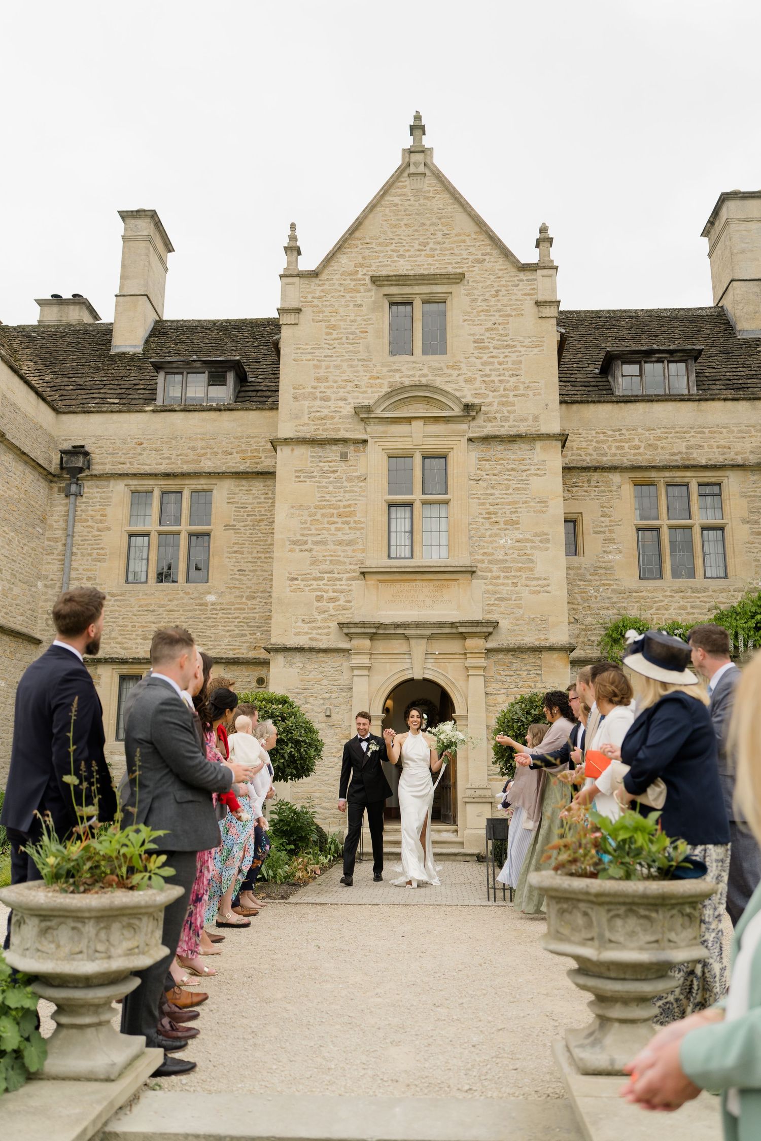 Bride and groom walk down outdoor aisle at elegant stone manor house wedding venue with guests lining the path.