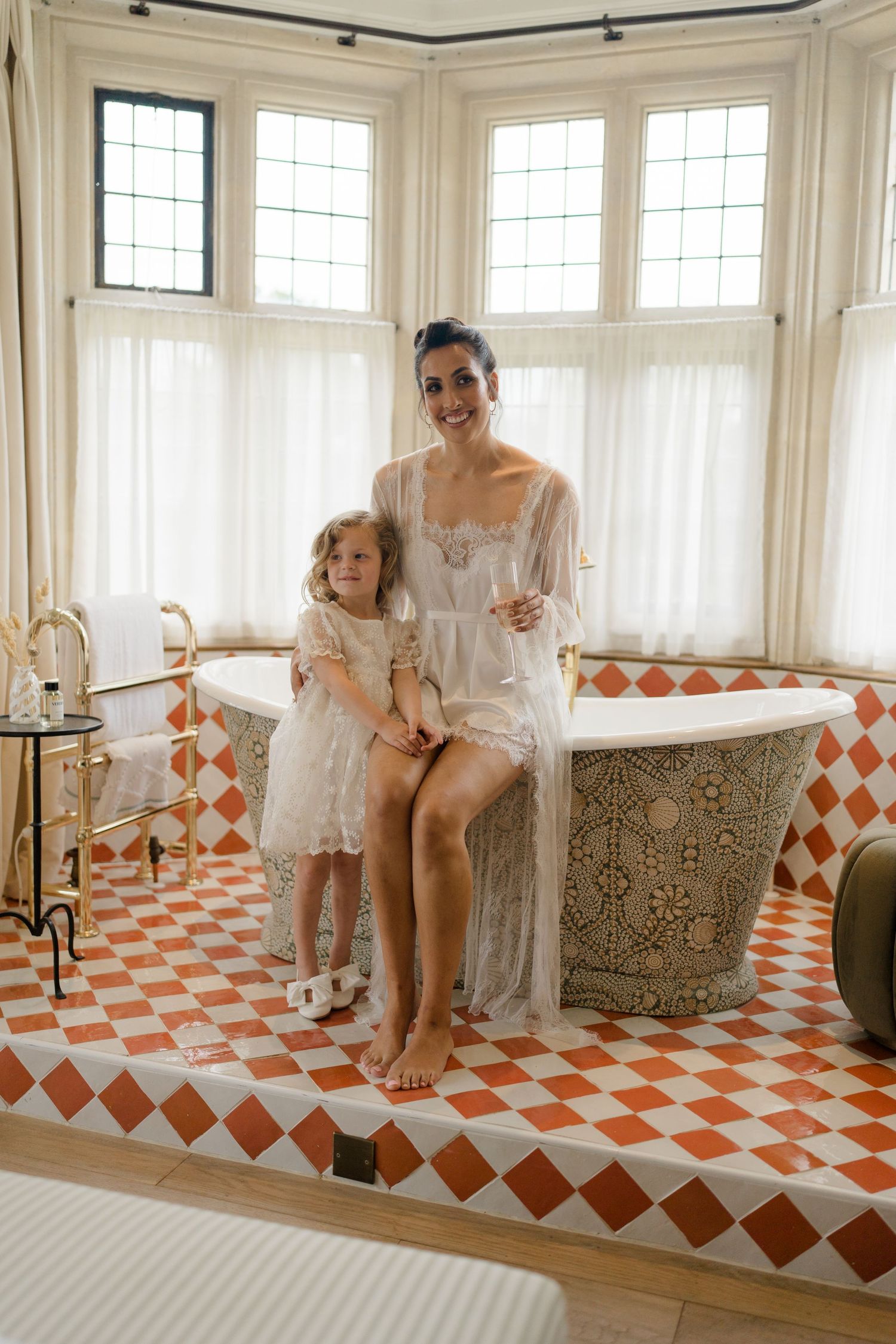 Bride in white lace robe poses with flower girl in ornate bathroom with red diamond tile floor.