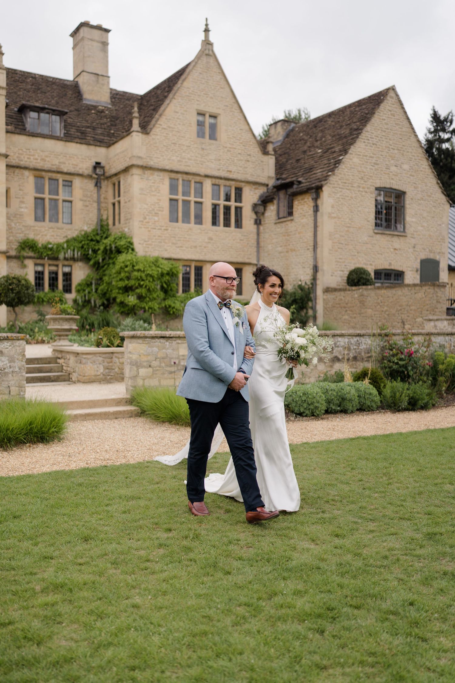 Bride and groom walk hand in hand on lawn before a Cotswolds manor house wedding venue.