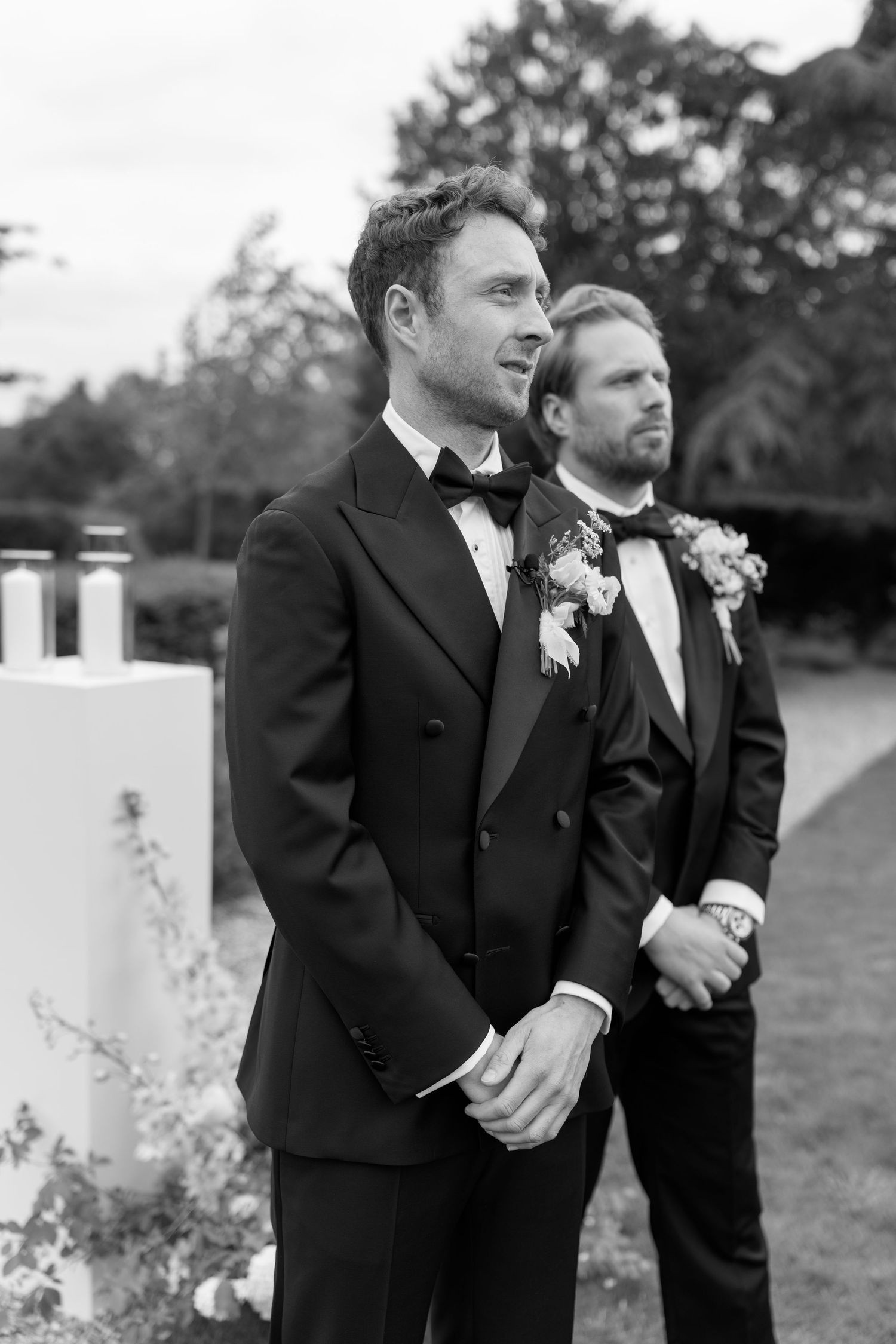 Groom in black tuxedo with boutonniere awaits bride at outdoor wedding ceremony with best man beside him.