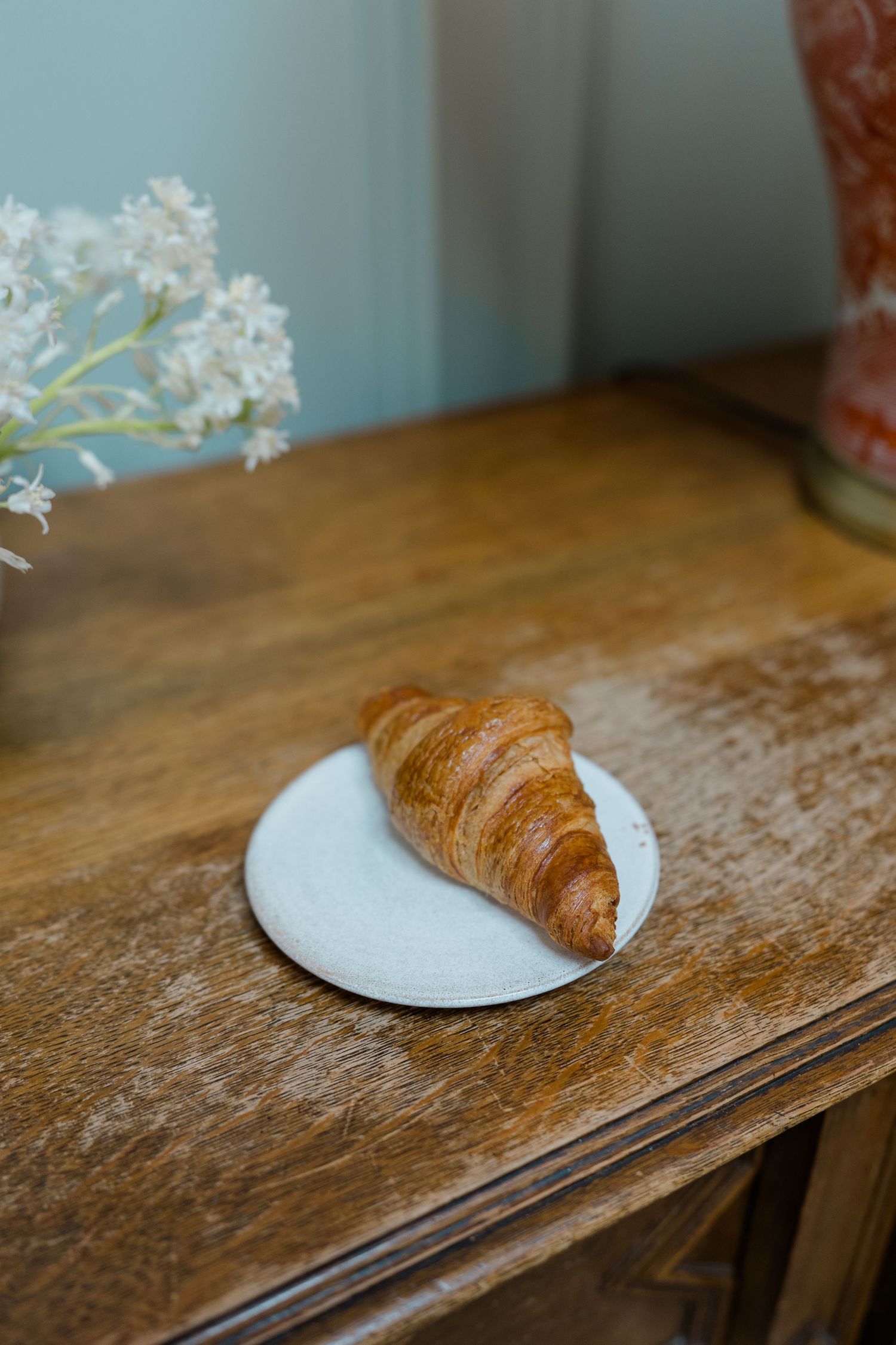 Golden croissant on a white plate atop a rustic wooden sideboard with flowers nearby.