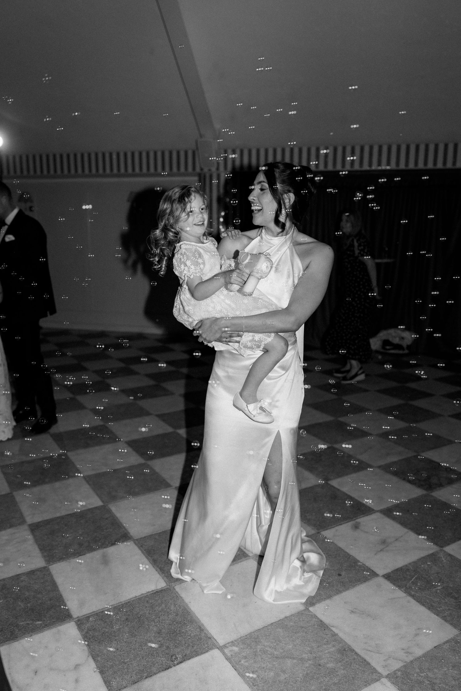 Bride dances joyfully while holding a young girl on a checkered dance floor at her reception.