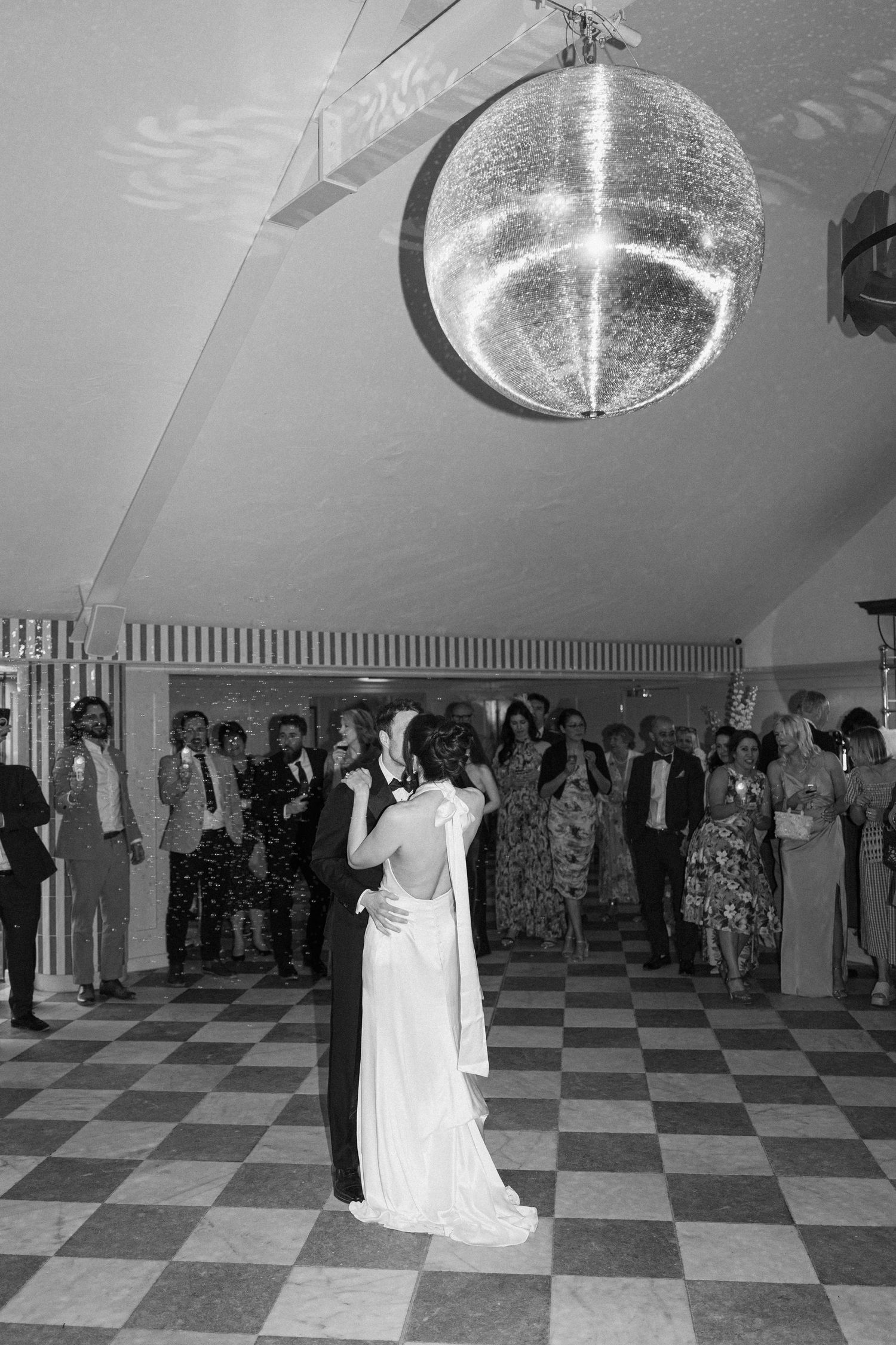 Bride and groom share first dance under sparkling disco ball on checkered dance floor in black and white.