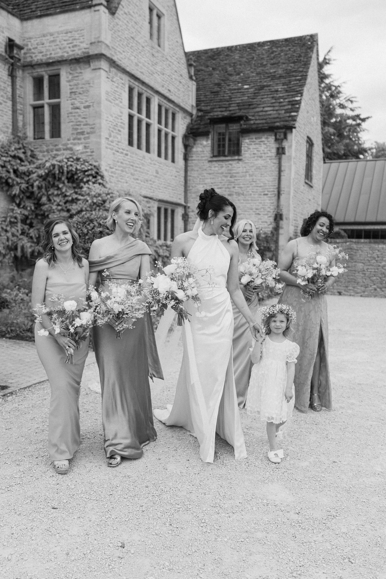 Black and white photo of bride with bridesmaids in elegant gowns and flower girl outside stone manor.