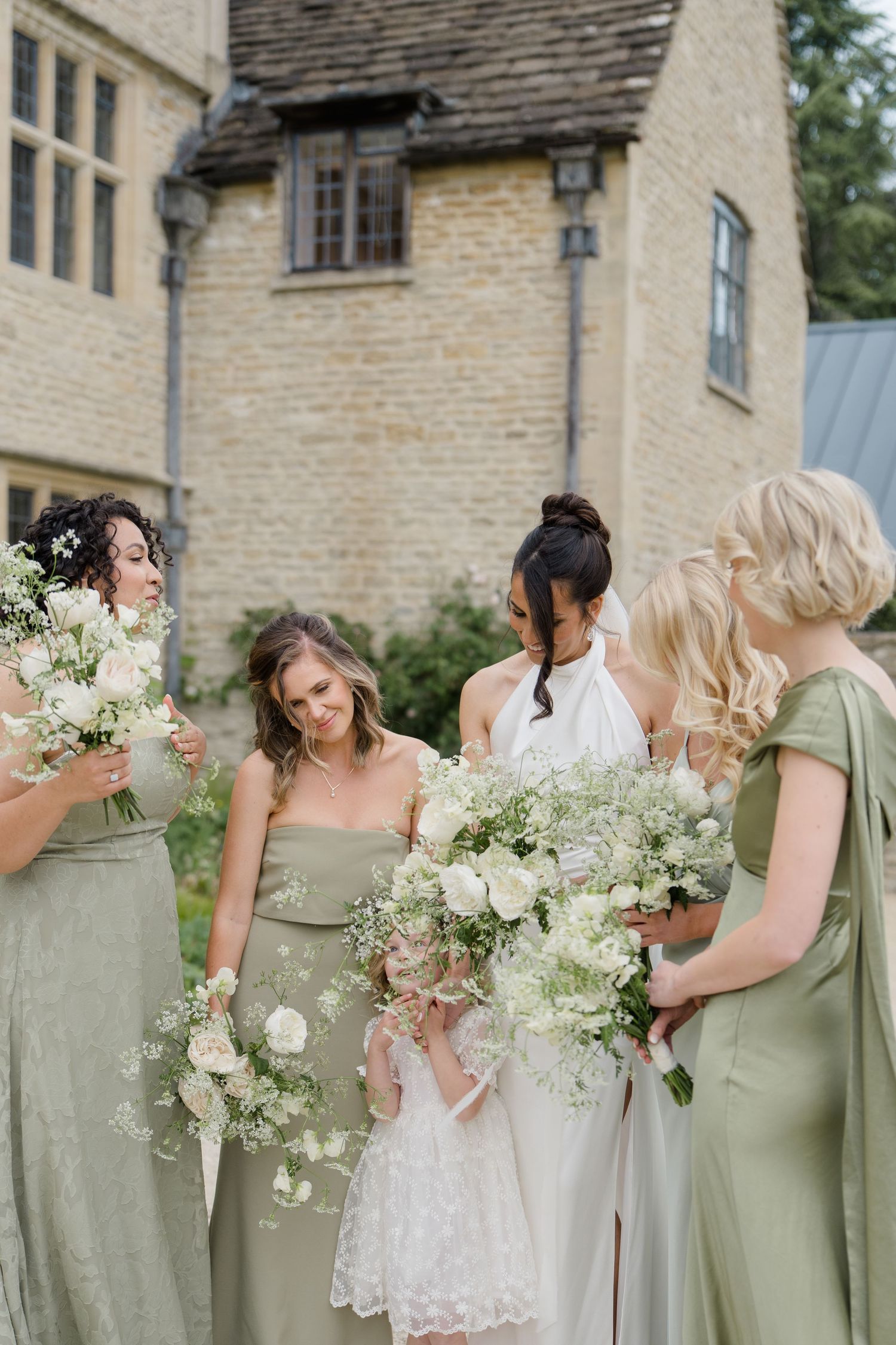 Bride and bridesmaids in sage green dresses admire white floral bouquets outside stone manor.