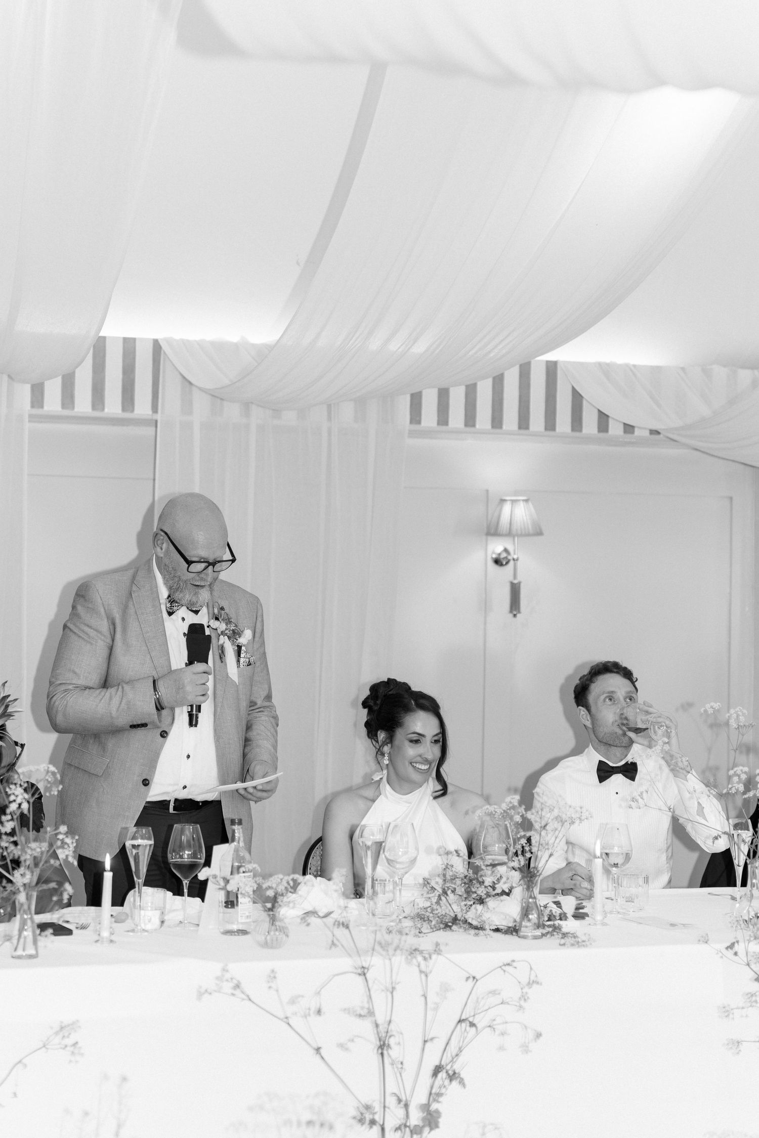 Father of bride giving speech while couple sit at top table in marquee reception.