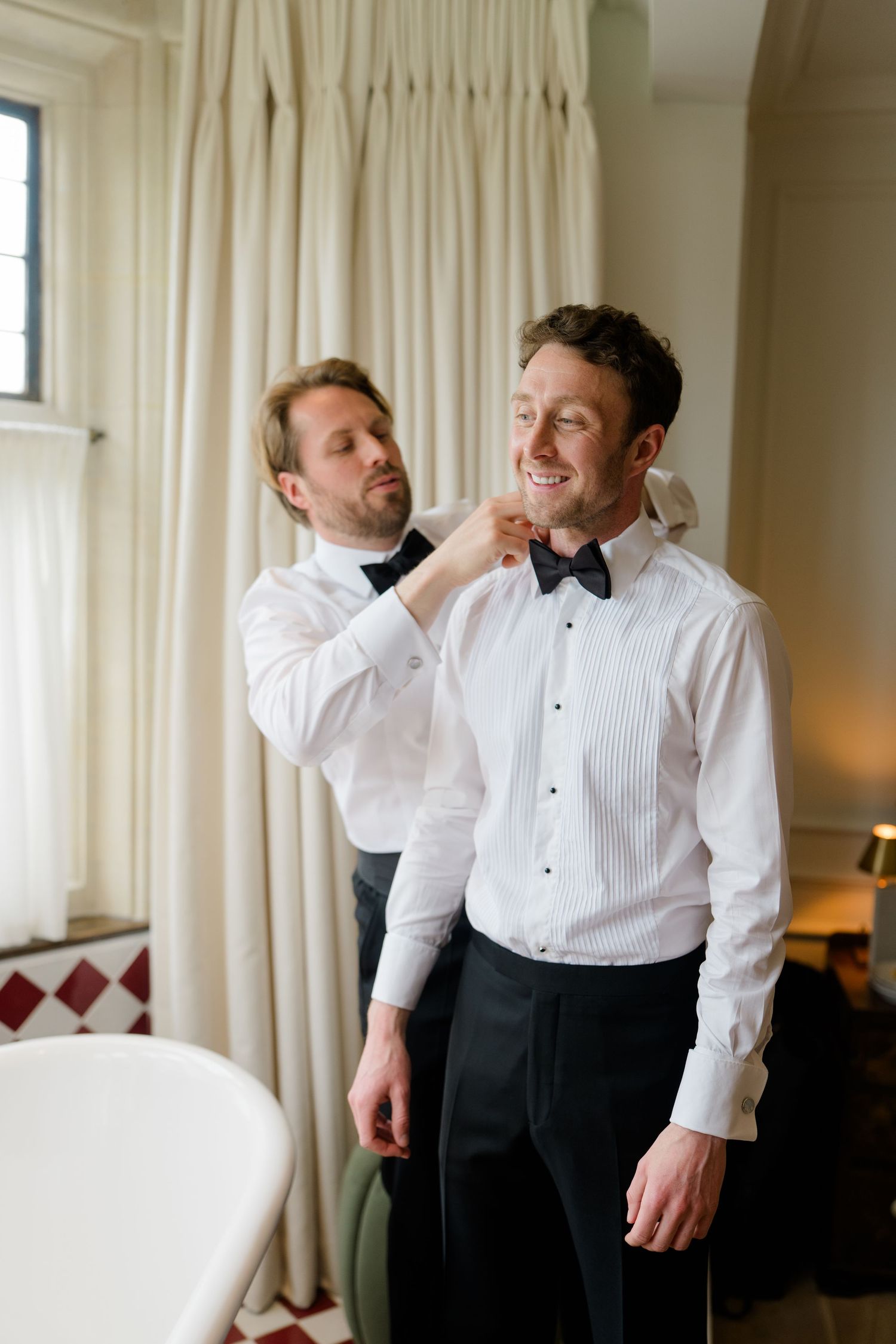 Groomsman helping groom adjust bow tie while getting dressed for wedding ceremony.