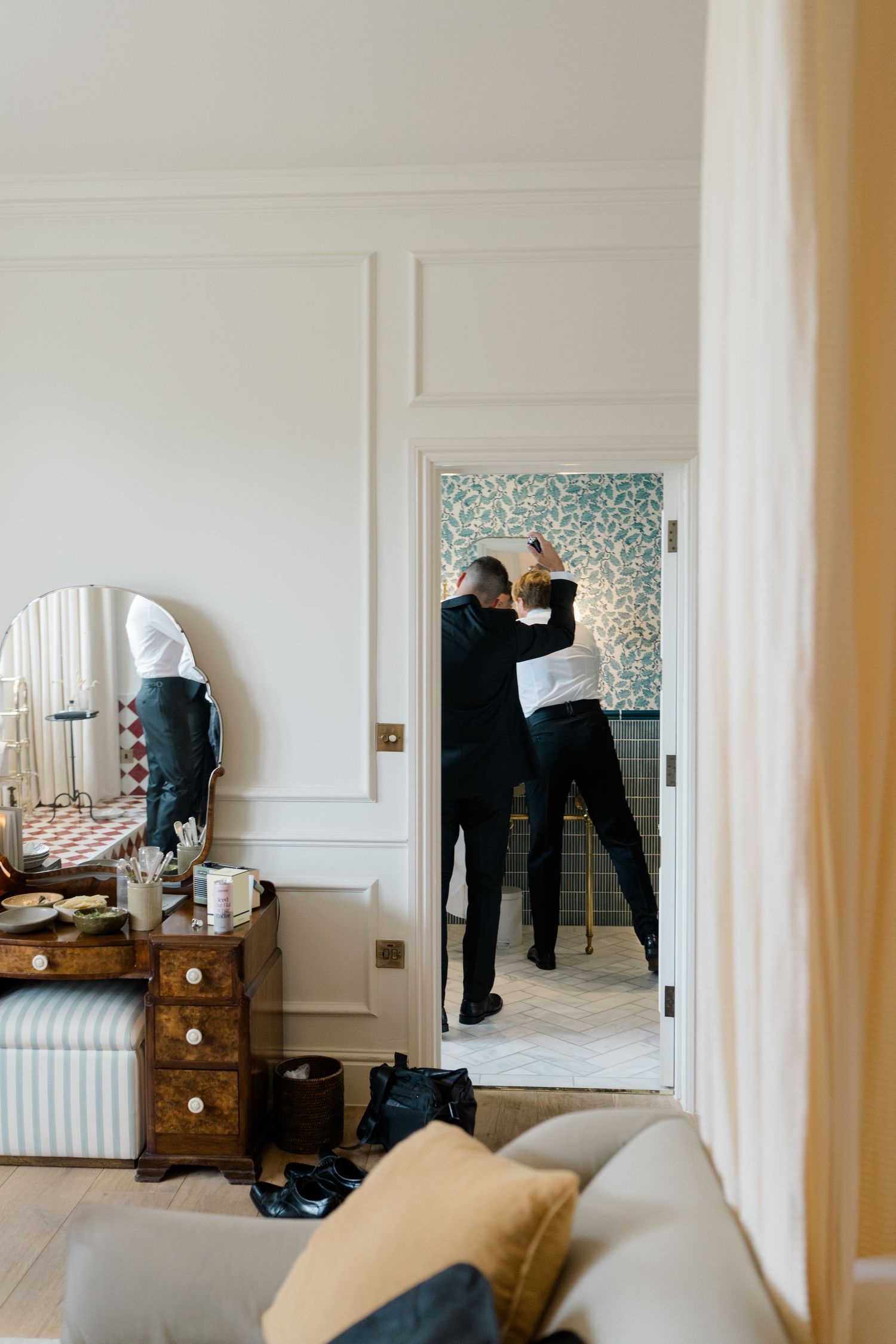 Groom getting ready with groomsman adjusting tie in elegant hotel room doorway.