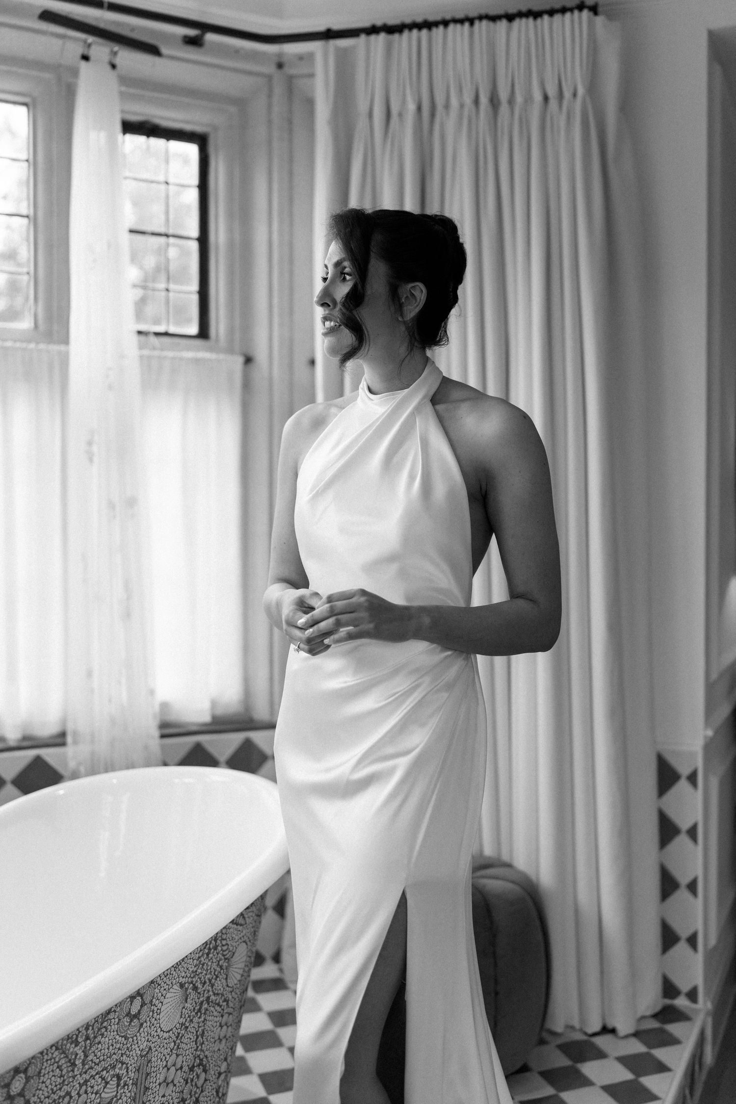 Black and white portrait of bride in halter neck wedding dress standing by a freestanding bathtub.