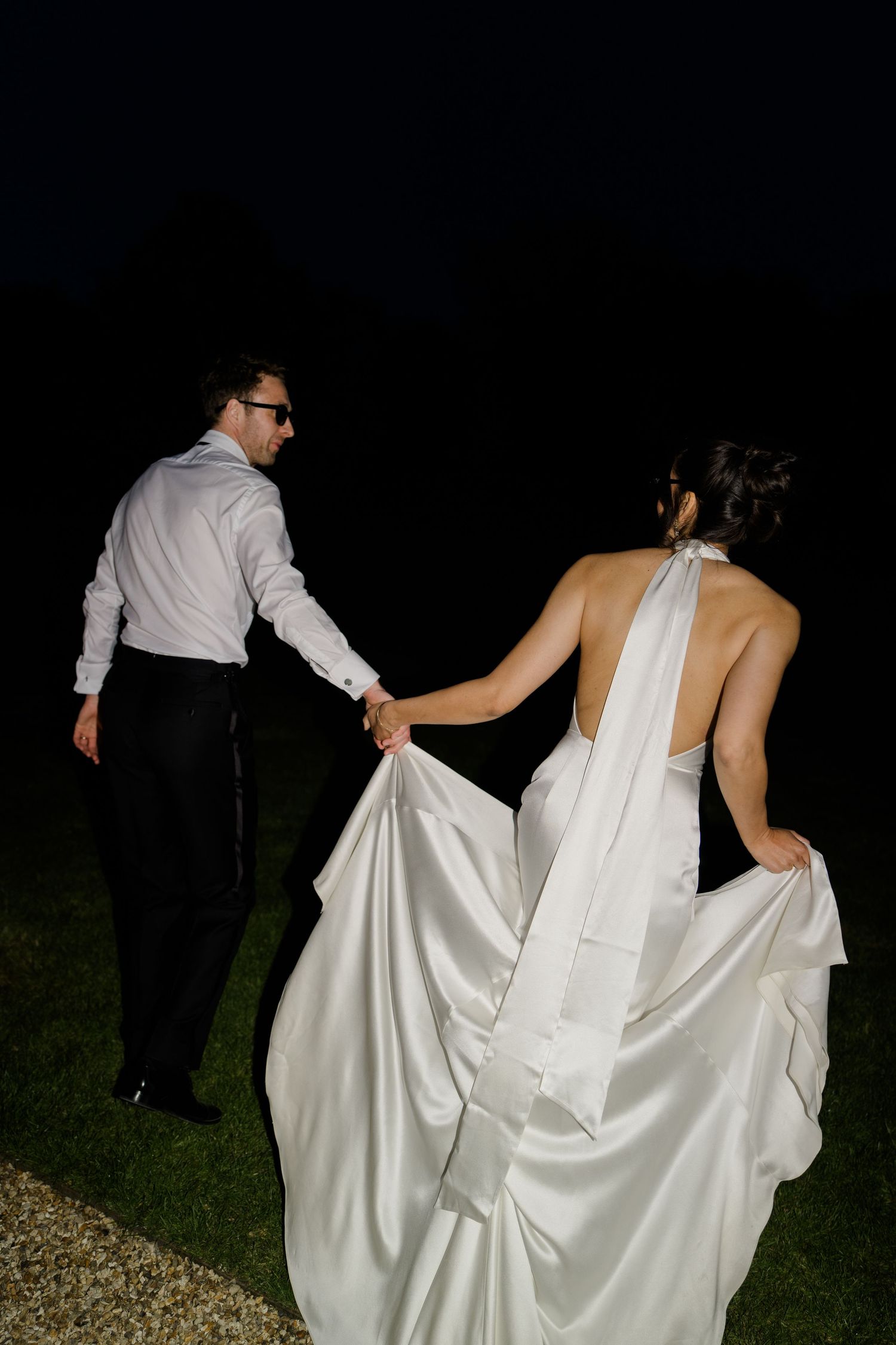 A bride in a white halter gown and groom hold hands walking through darkness at night.