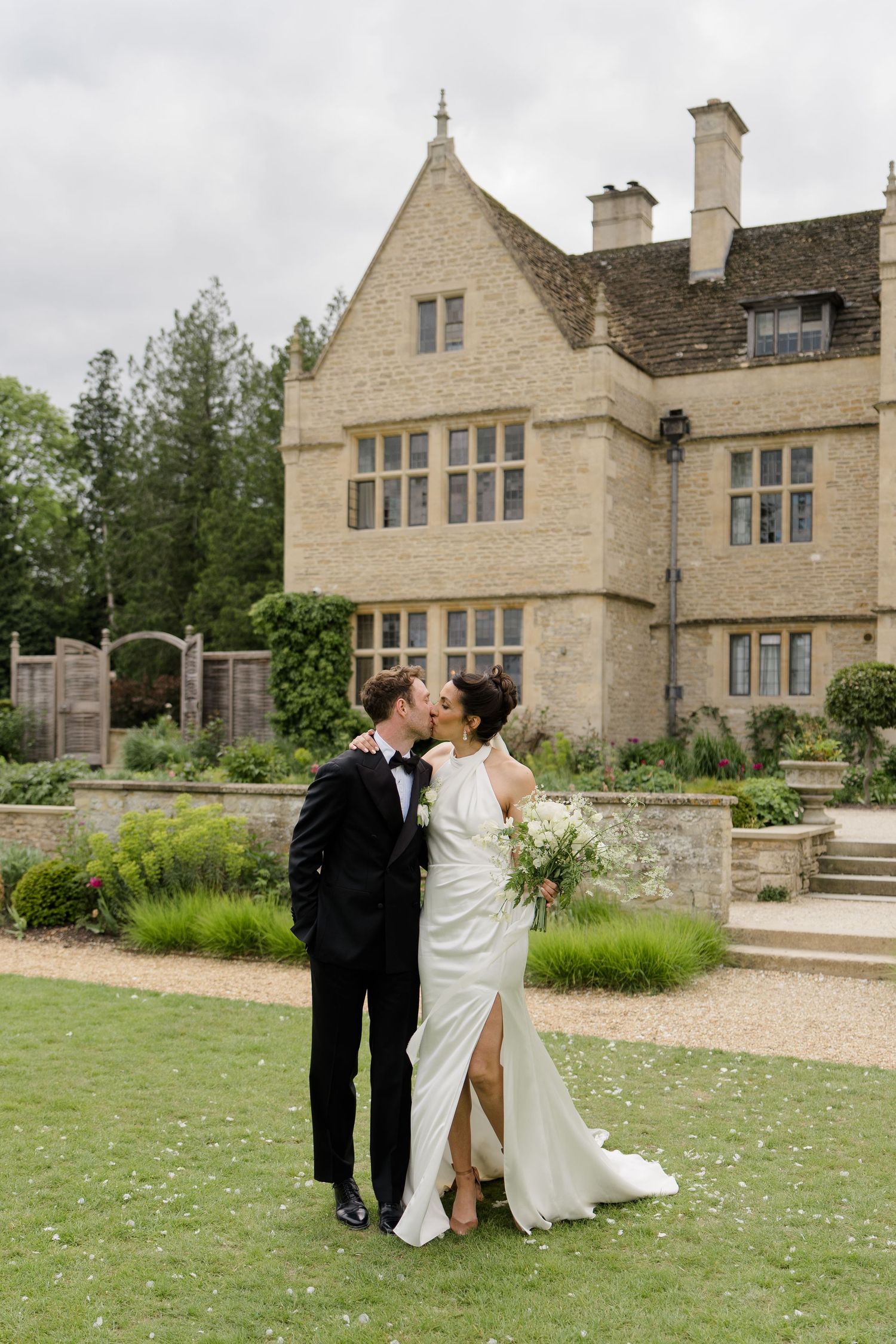 Bride and groom kiss on lawn in front of elegant stone manor house on wedding day.