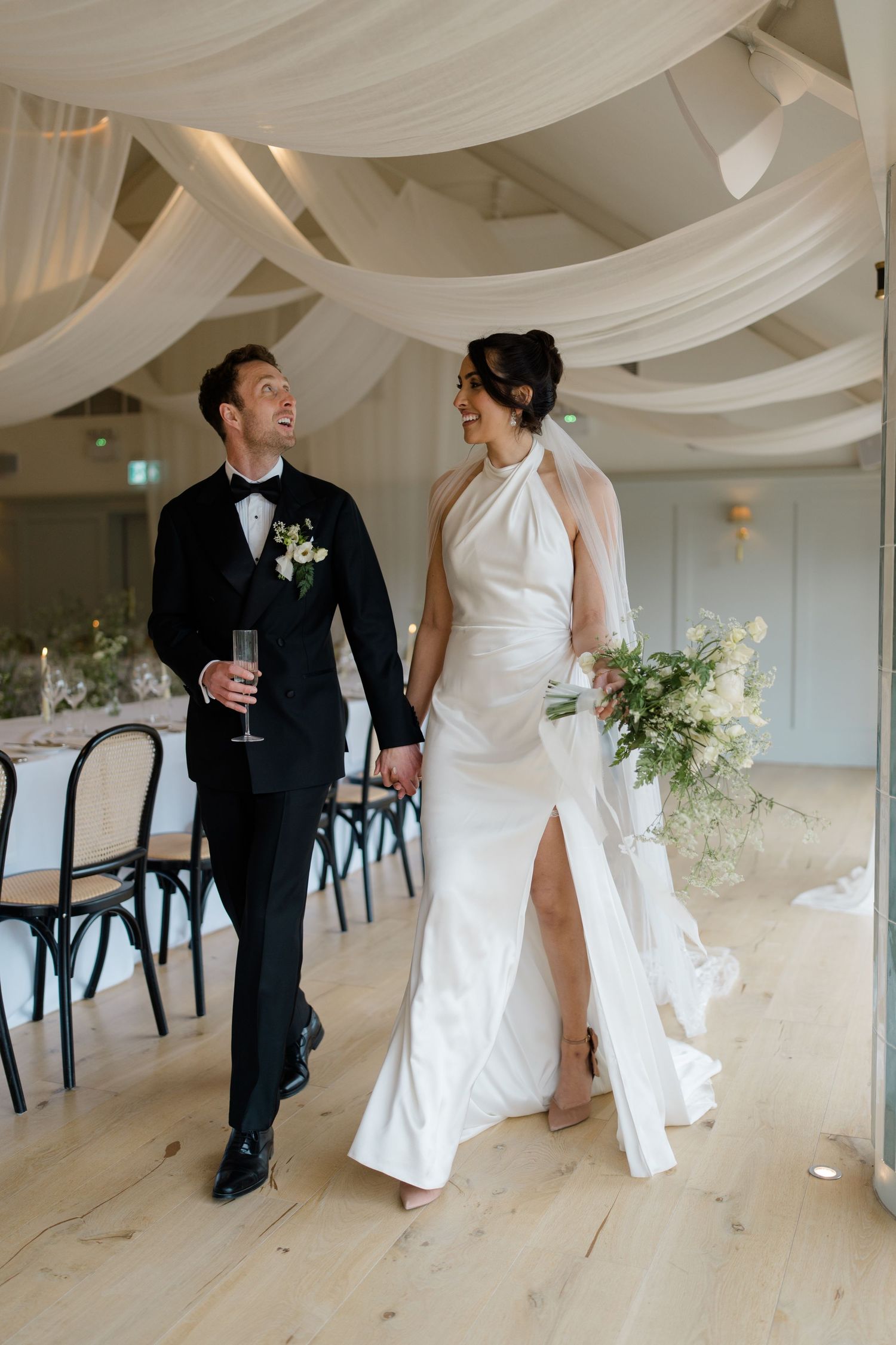 Bride and groom walk laughing through draped reception hall, bride in white halter gown with slit, groom in black tuxedo.