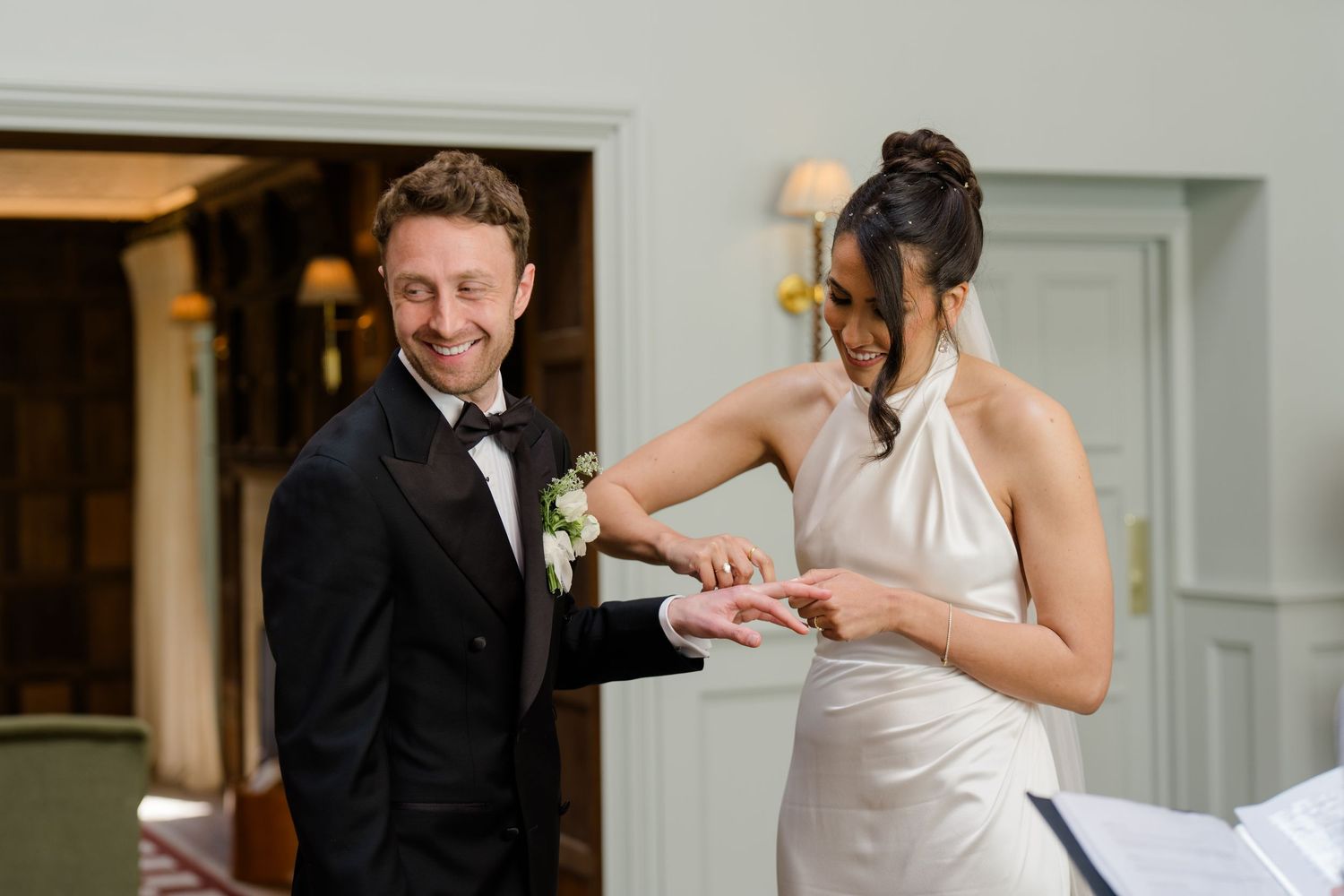 Groom in black tuxedo placing a ring on his smiling bride's finger during an intimate wedding ceremony.