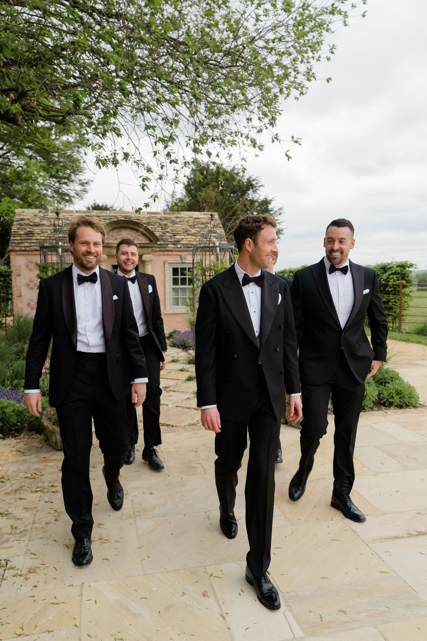 Four groomsmen in black tuxedos and bow ties walk and laugh together at a Cotswolds wedding venue.