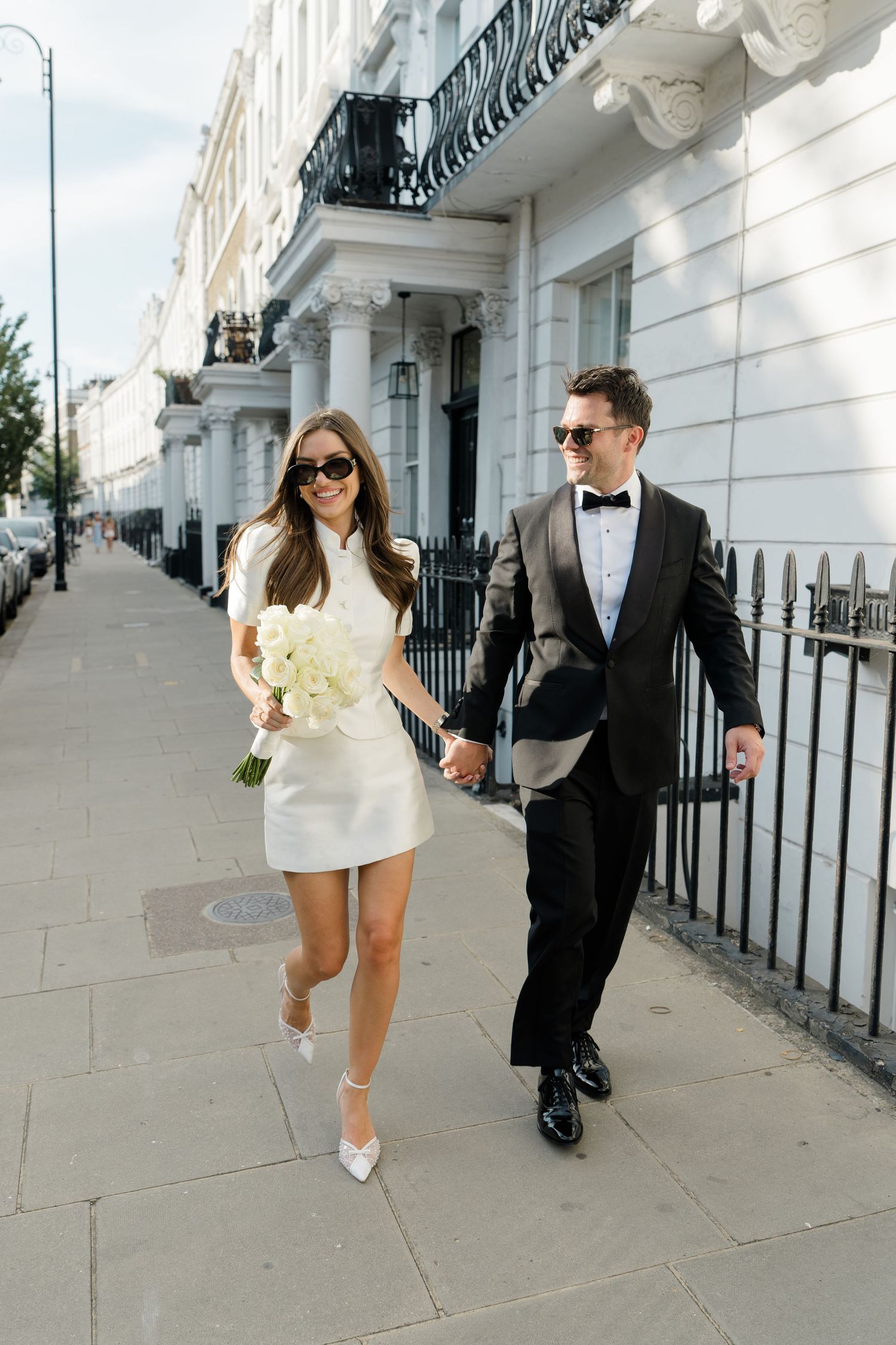 Stylish bride and groom holding hands walking along a white Georgian townhouse London street.