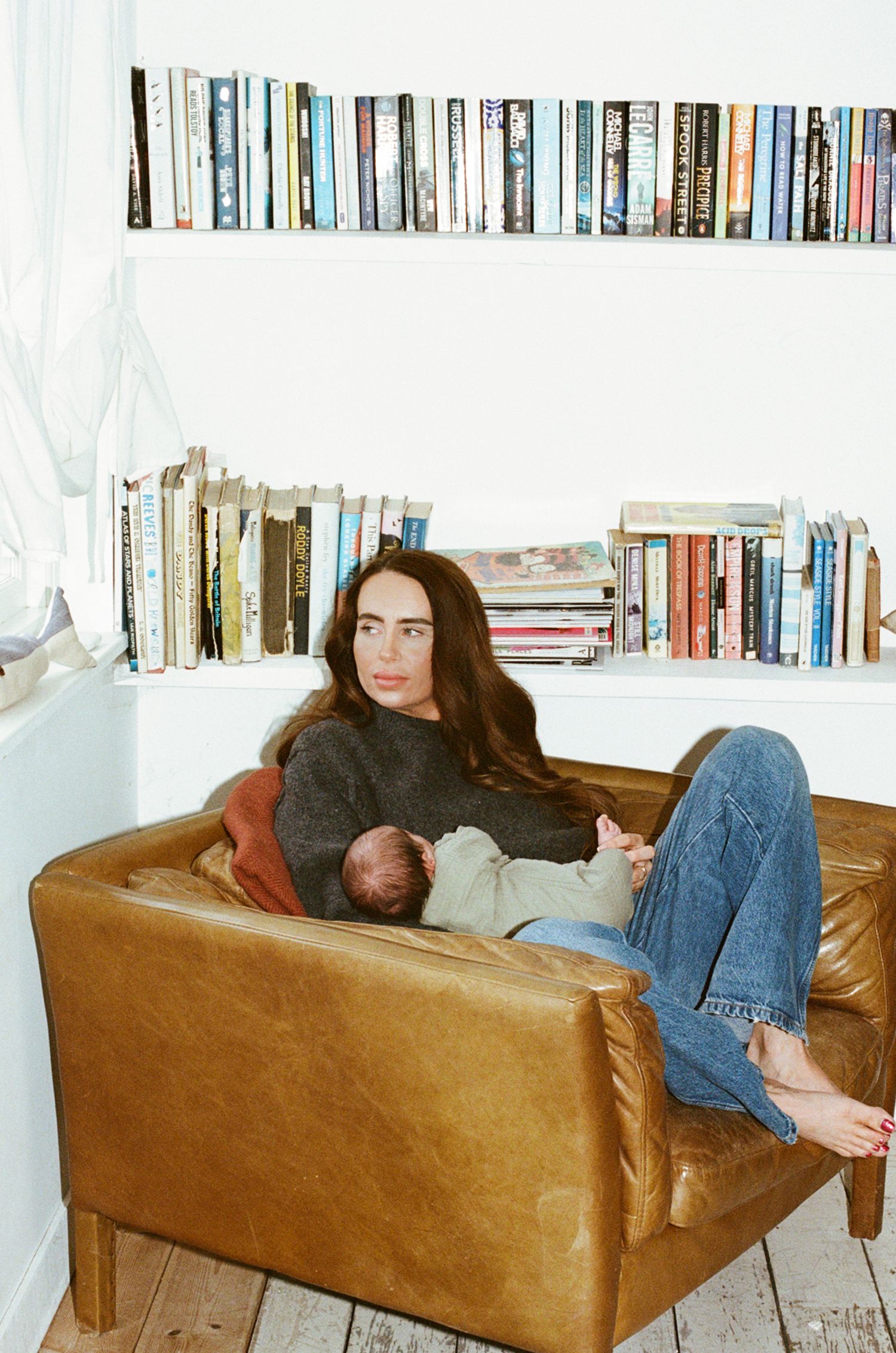 A woman breastfeeding a newborn on a tan leather armchair surrounded by bookshelves in a bright white room.