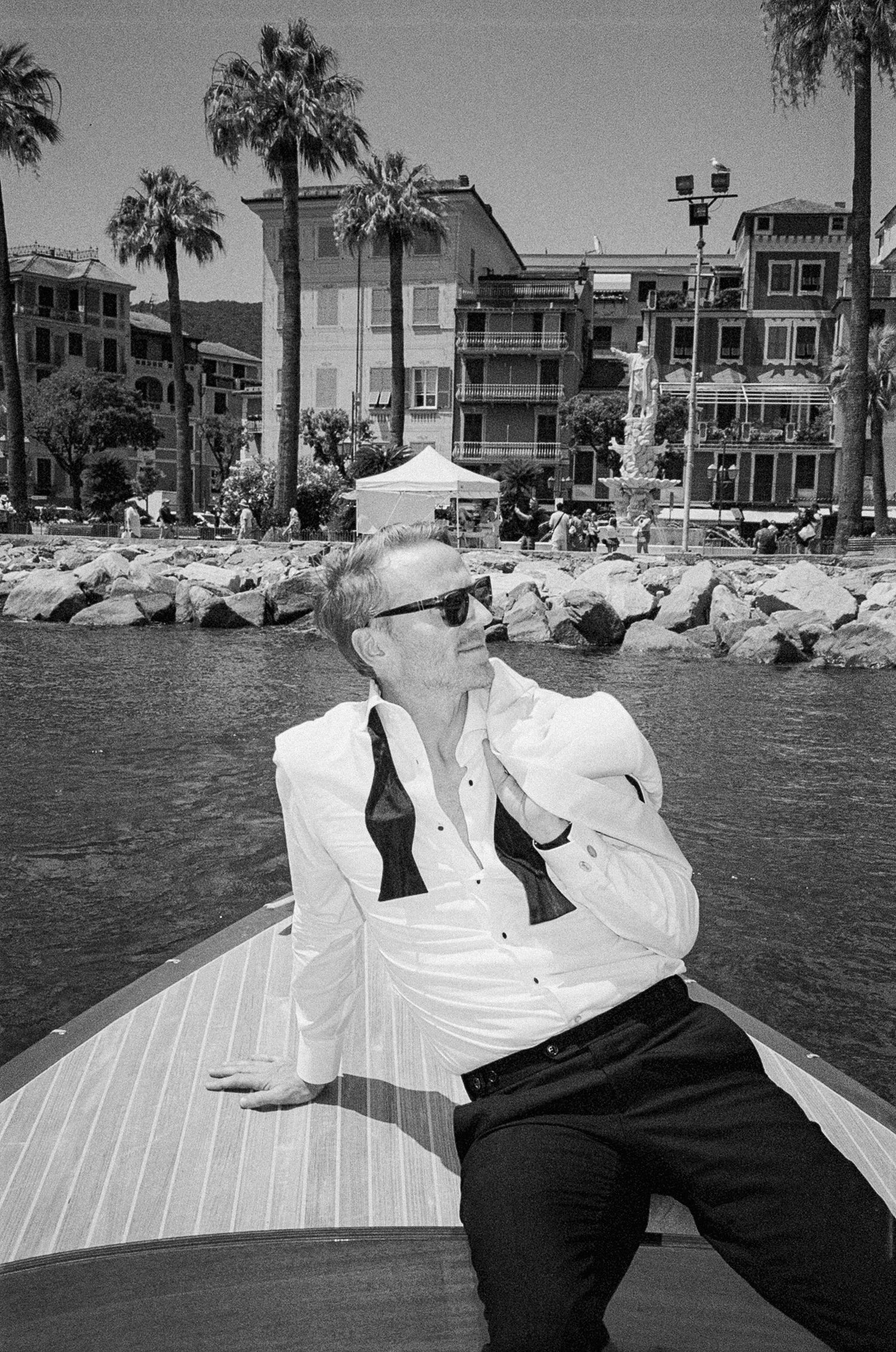 Man in white shirt and sunglasses relaxing on a boat near a Mediterranean waterfront, black and white photo.