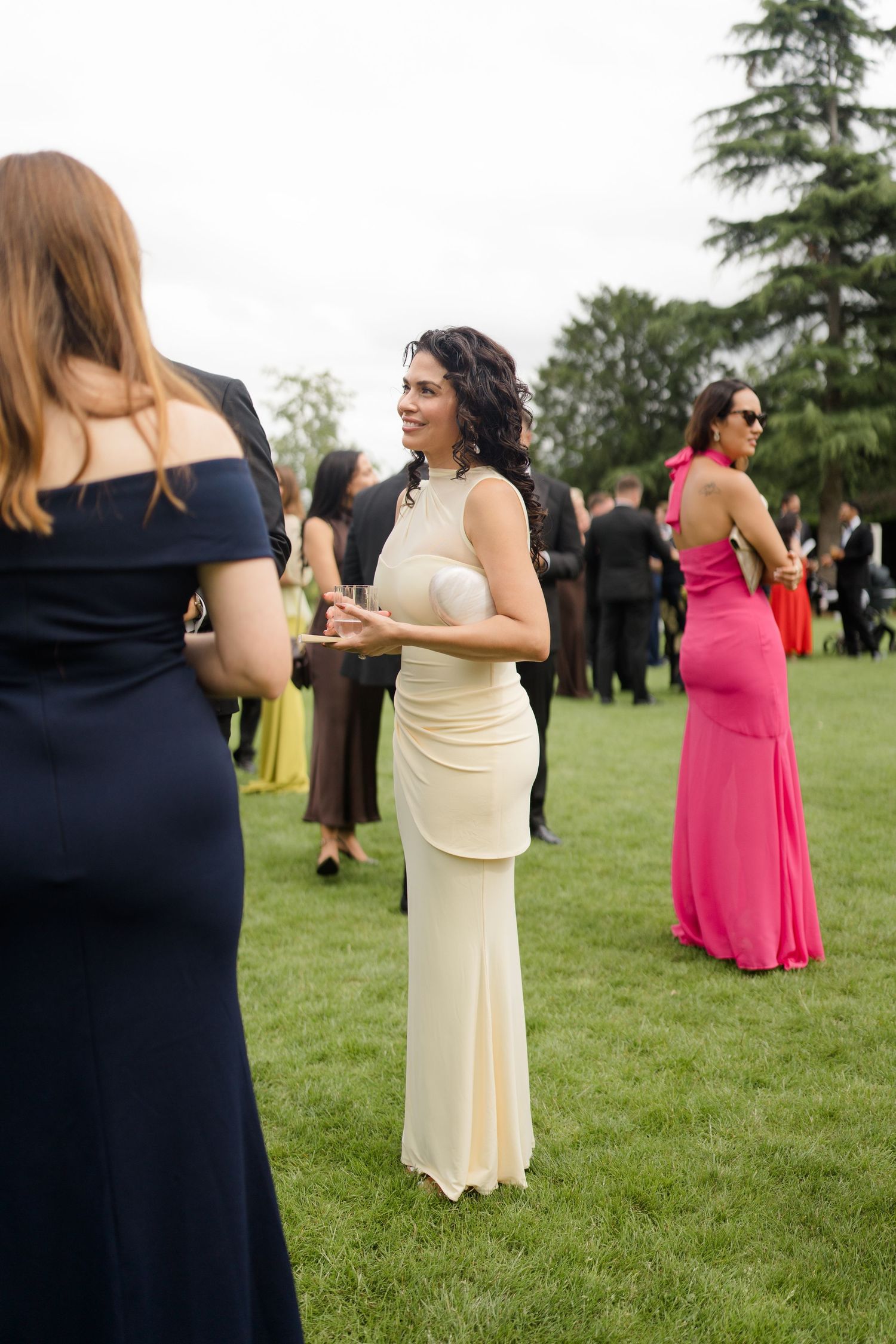 Wedding guests mingling on lawn, woman in cream gown conversing with others in formal attire outdoors.