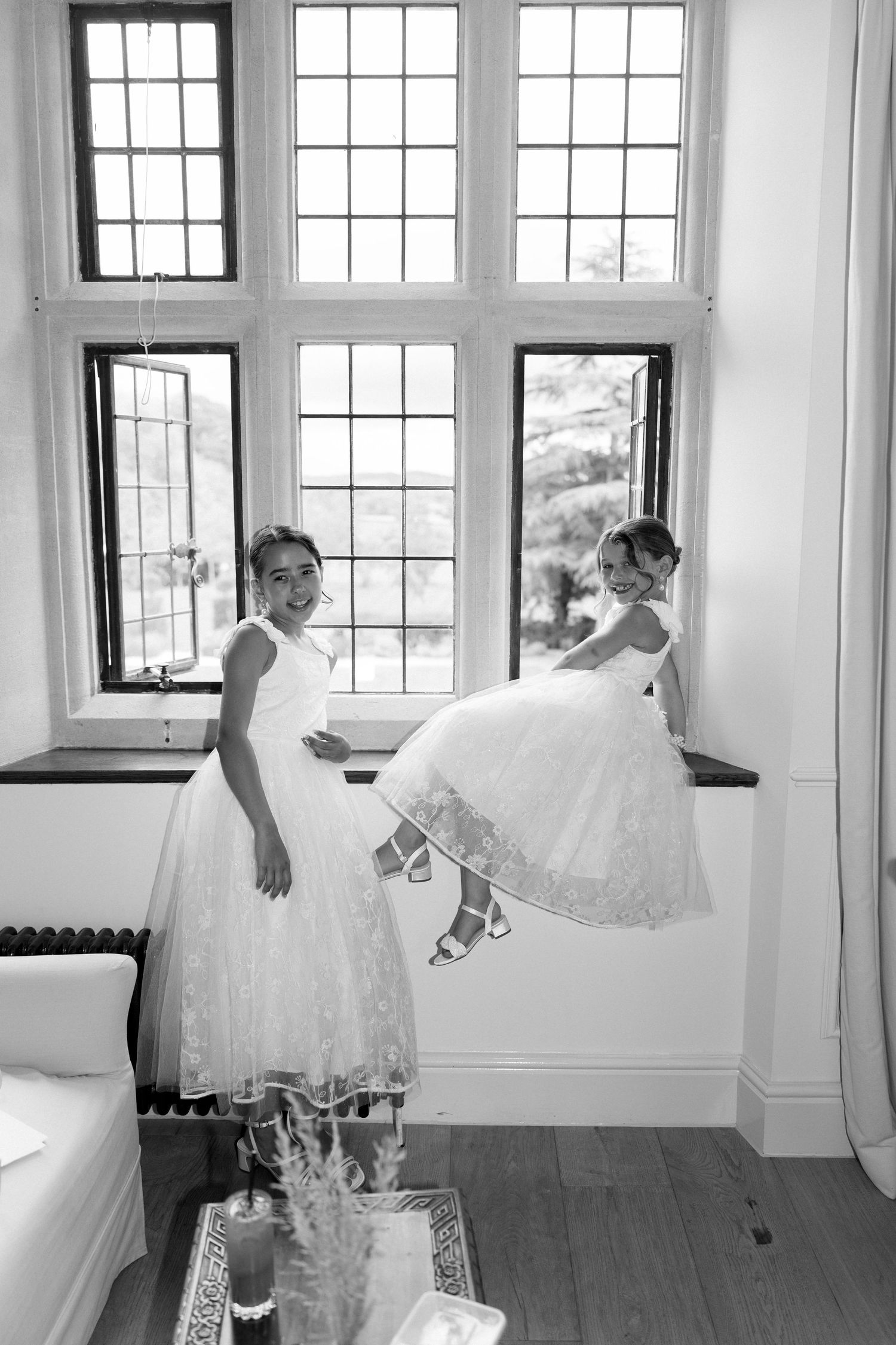 Two flower girls in white lace dresses posing by large leaded glass windows, black and white photo.