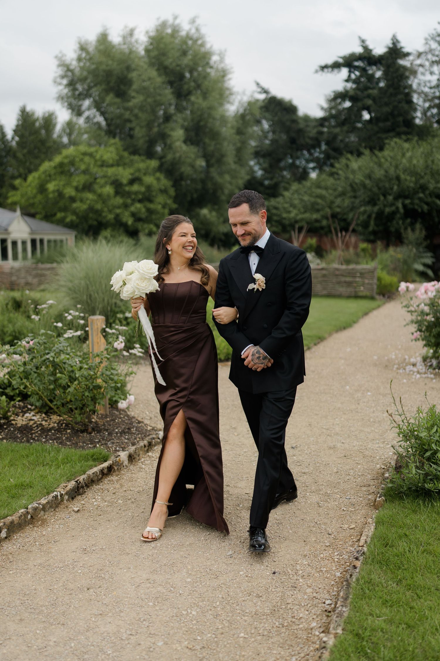 Smiling bridesmaid in chocolate satin gown with slit and groomsman in black tuxedo stroll through garden wedding path.