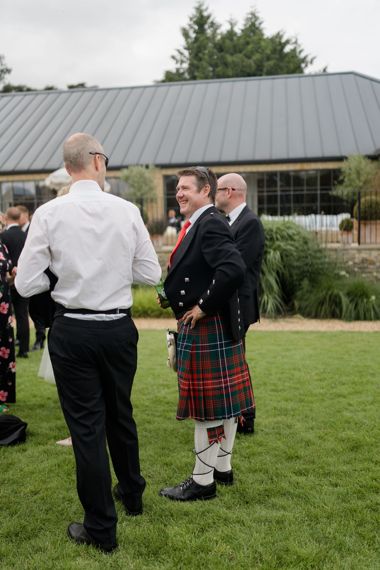 Wedding guest in traditional Scottish kilt with red tartan laughing with guests outdoors.
