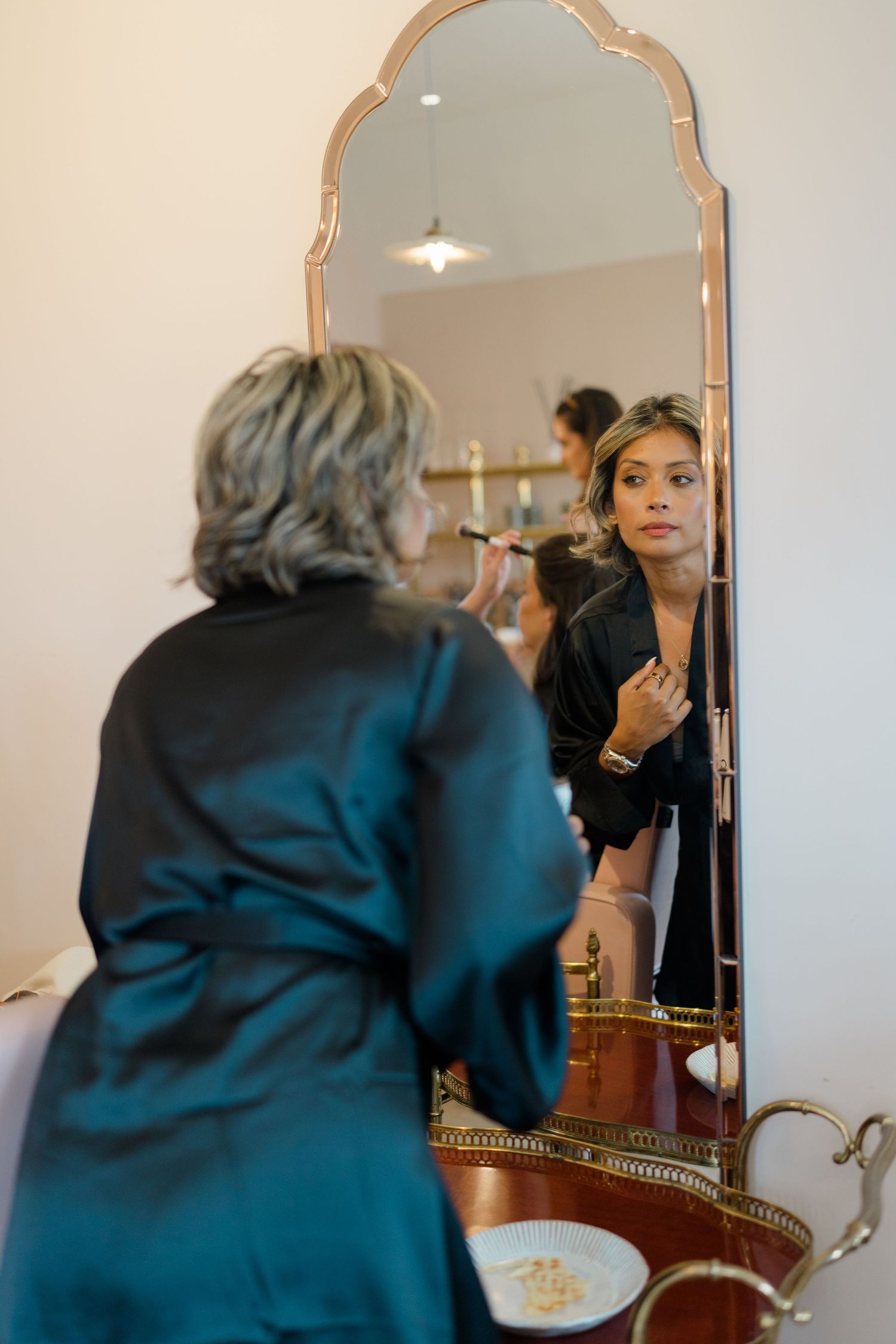Bride applies makeup while reflected in ornate gold mirror during wedding preparations.